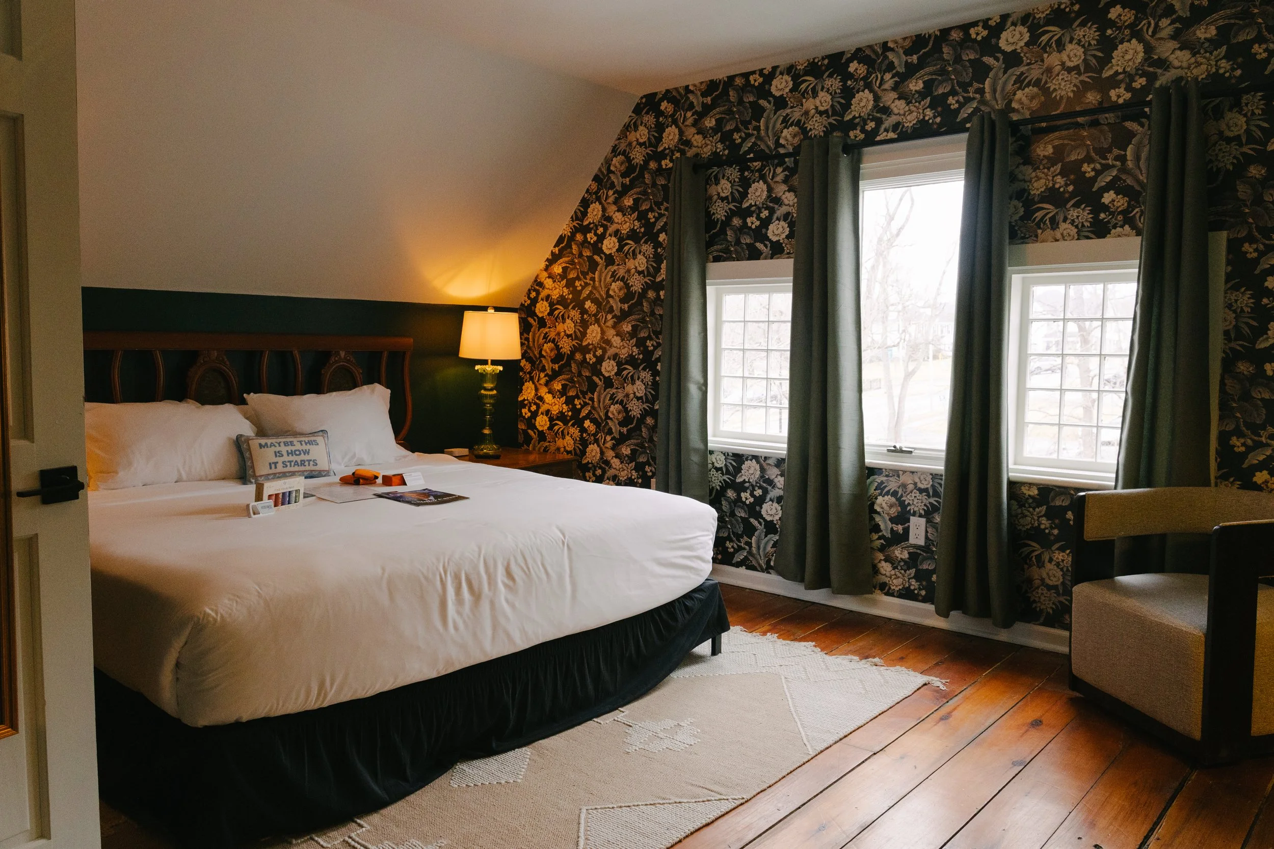 A cozy hotel room with a large bed, a wooden headboard, a bedside lamp, and a decorative dark floral wallpaper. There are three windows with green curtains, a small seating area with an armchair, and a beige rug on wooden floors.