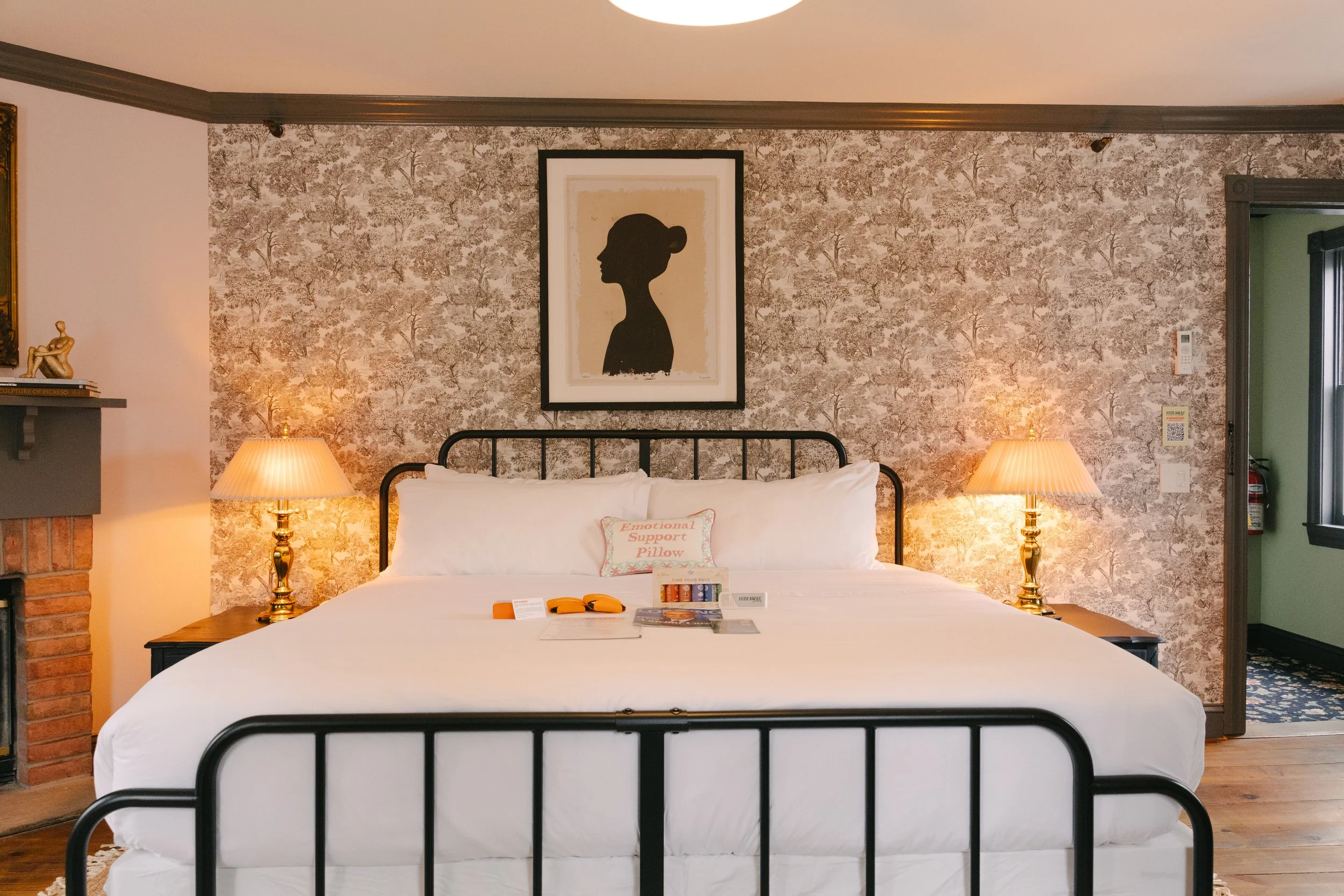 A bedroom with a metal bed frame, white bedding, and pillows. Two bedside lamps on wooden nightstands. Framed silhouette artwork of a woman on the wall. Decorative wallpaper behind the bed. A pillow labeled 'Emotional Support Pillow' and reading mate