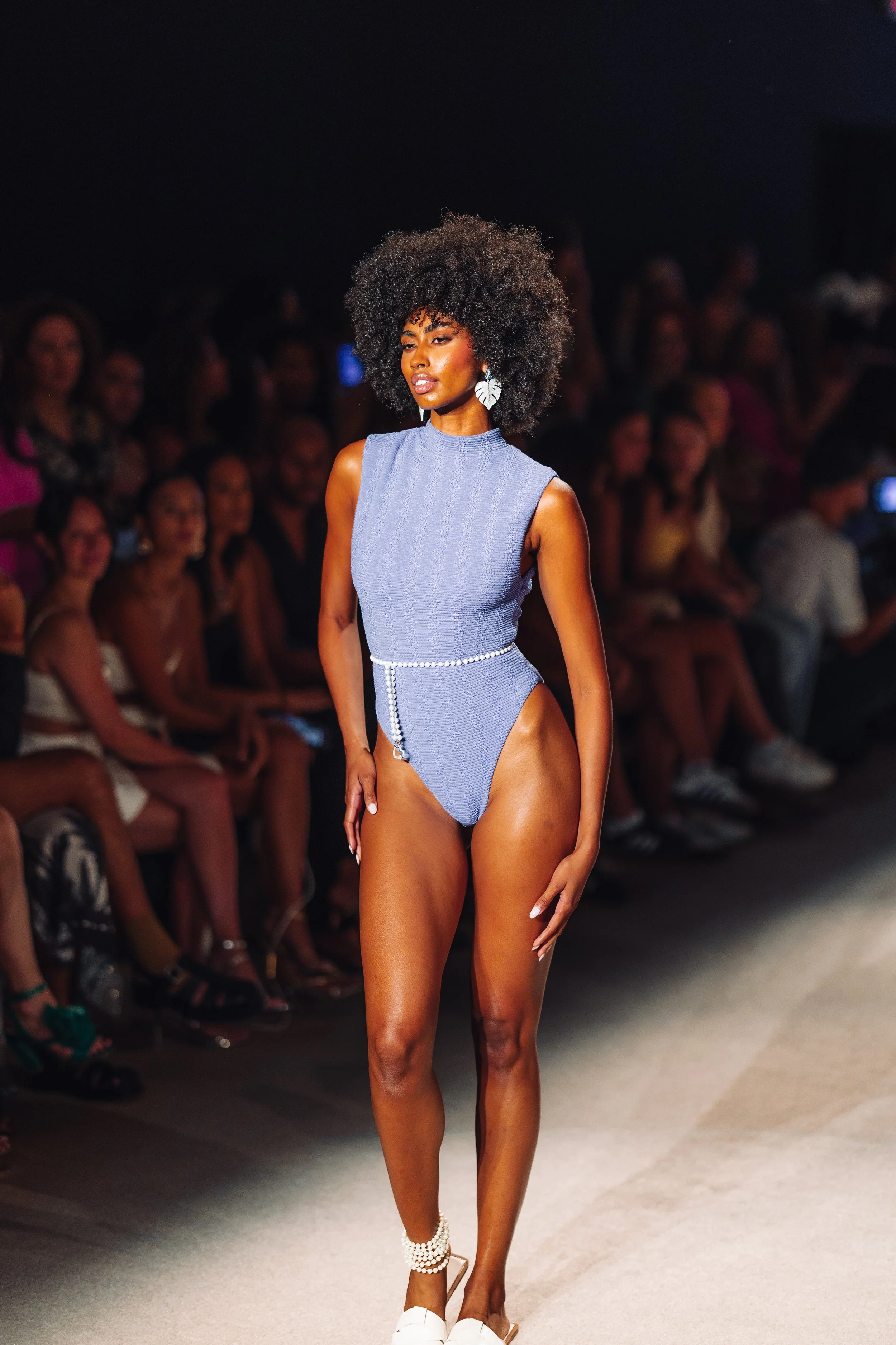 A model walking on a runway wearing a sleeveless, high-neck, lavender bodysuit with a pearl waist accessory in front of an audience.