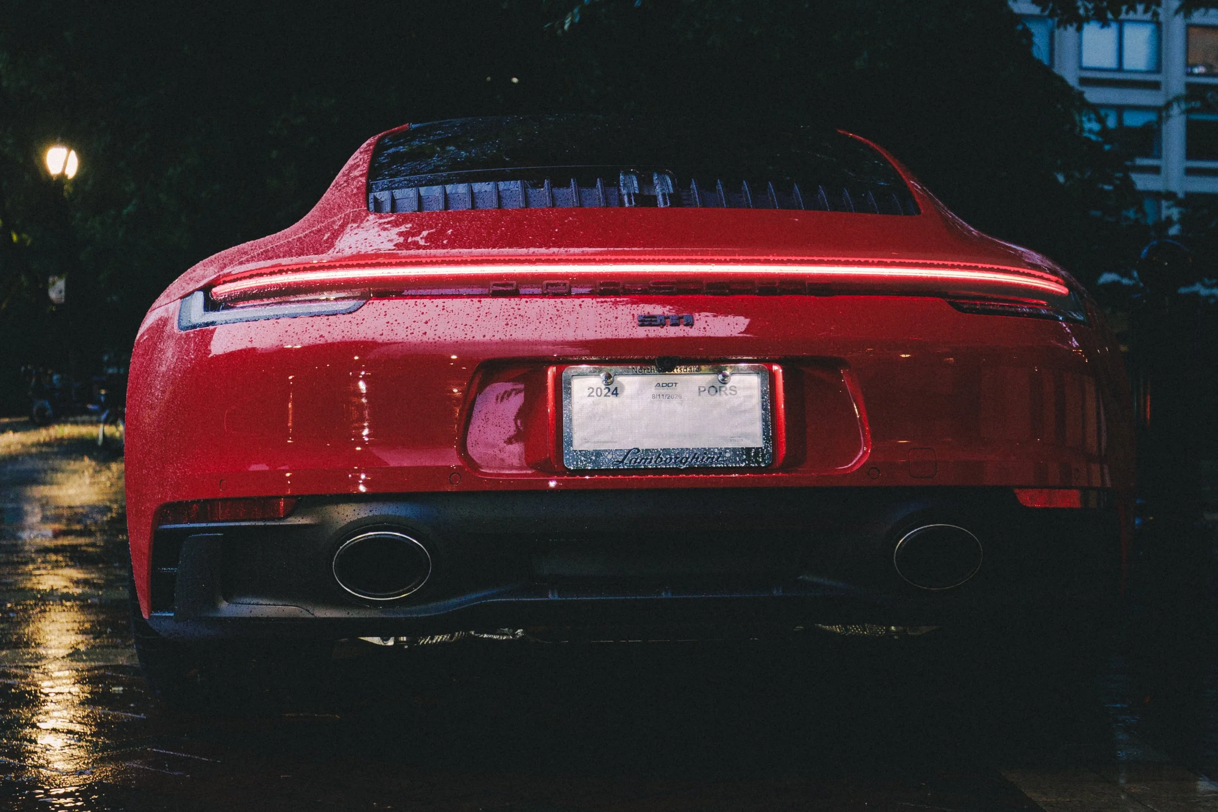 Red sports car, possibly a Porsche, parked at night with wet surface reflecting light, rain droplets on the car, rear view showing dual exhausts, a sleek design, and a rear spoiler.