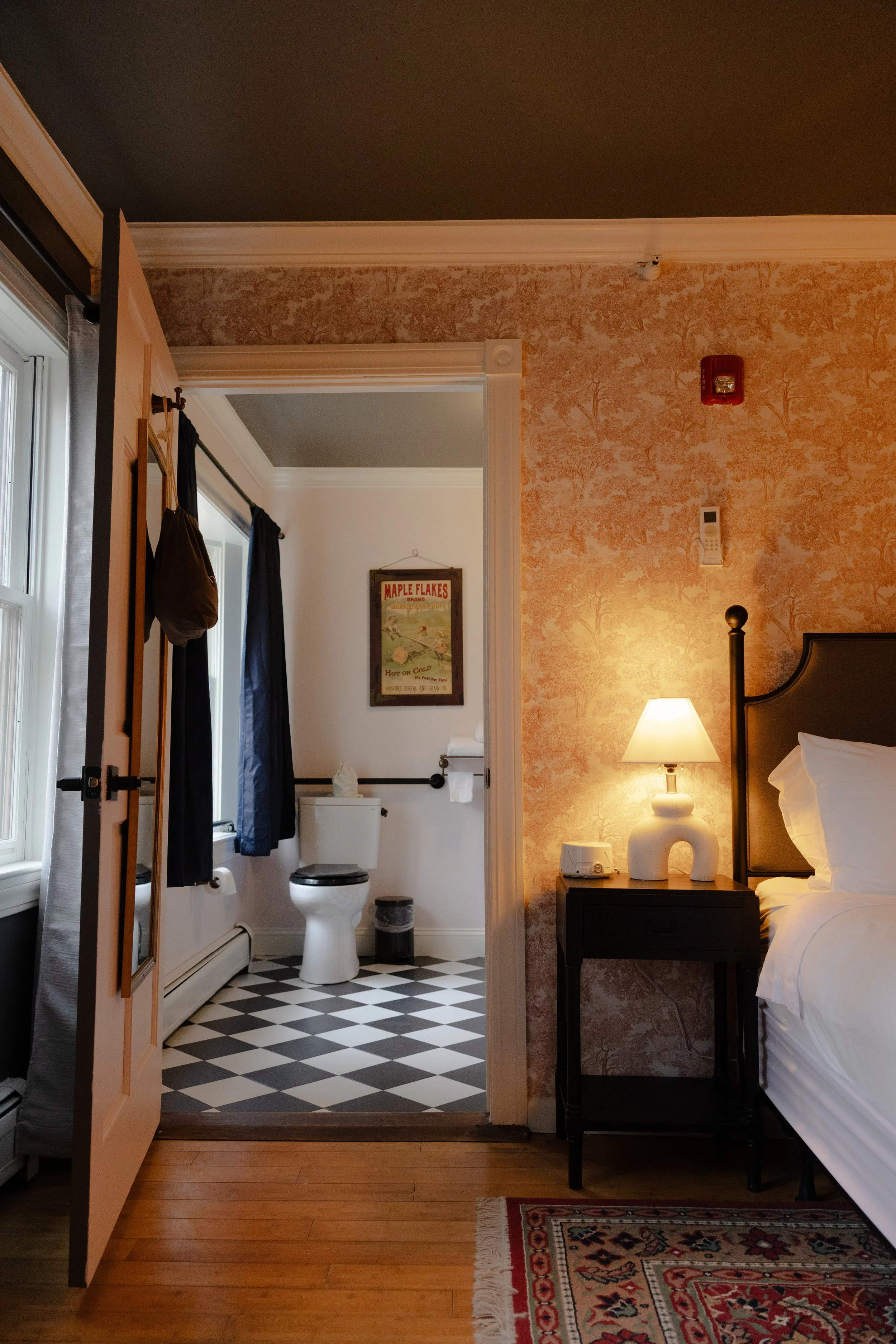 A bedroom with orange patterned wallpaper, a bedside table with a lamp, and a view into a bathroom with black and white checkered floor, toilet, and framed vintage Maple Flakes advertisement.