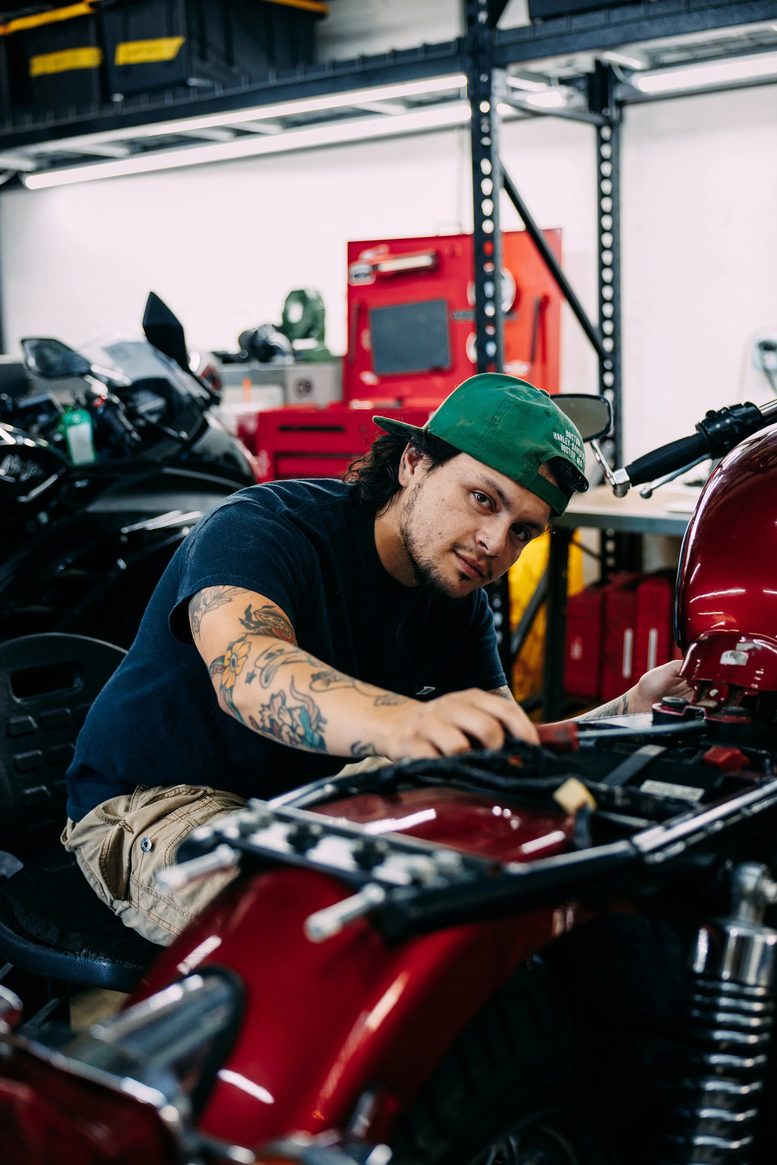 A man with tattoos and a green cap working on a motorcycle in a garage with tools and equipment in the background.
