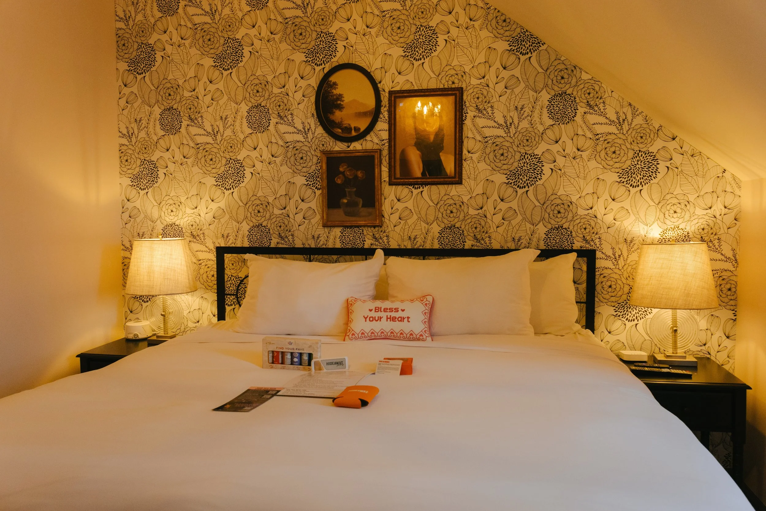 A cozy hotel bedroom with a large bed, white bedding, and decorative pillows. Two bedside lamps illuminate the room. The wall behind the bed has floral patterned wallpaper with framed artwork, including a portrait and landscape scenes.