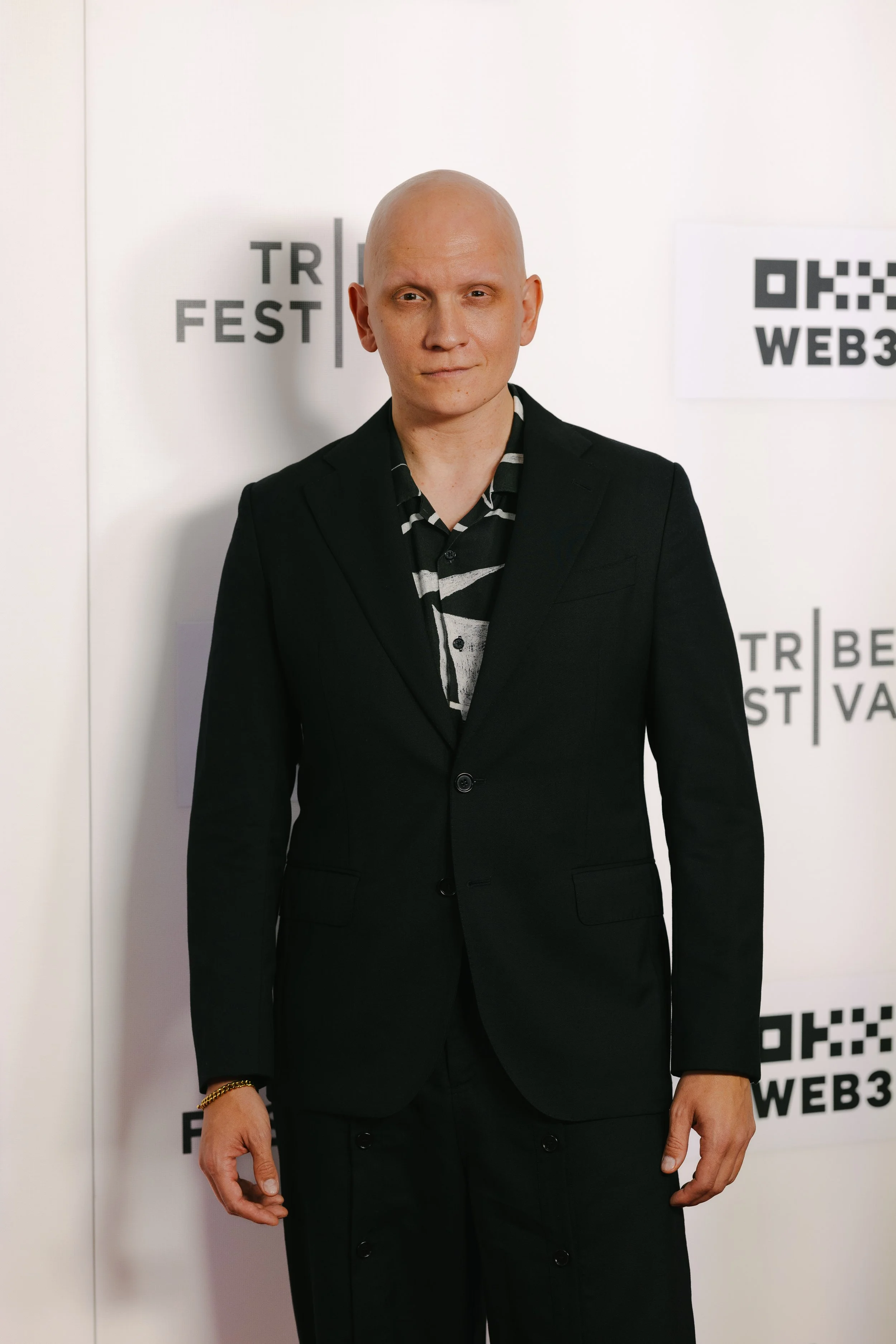 A person with a shaved head wearing a black suit and a black and white patterned shirt standing in front of a white backdrop with black text and QR codes.