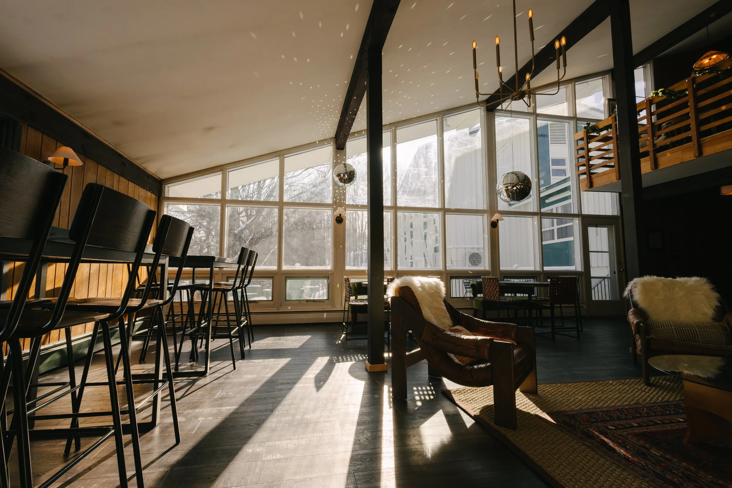 Sunlit interior of a cozy lounge with high windows, wooden walls, black chairs, leather armchairs with fluffy pillows, and hanging disco balls.
