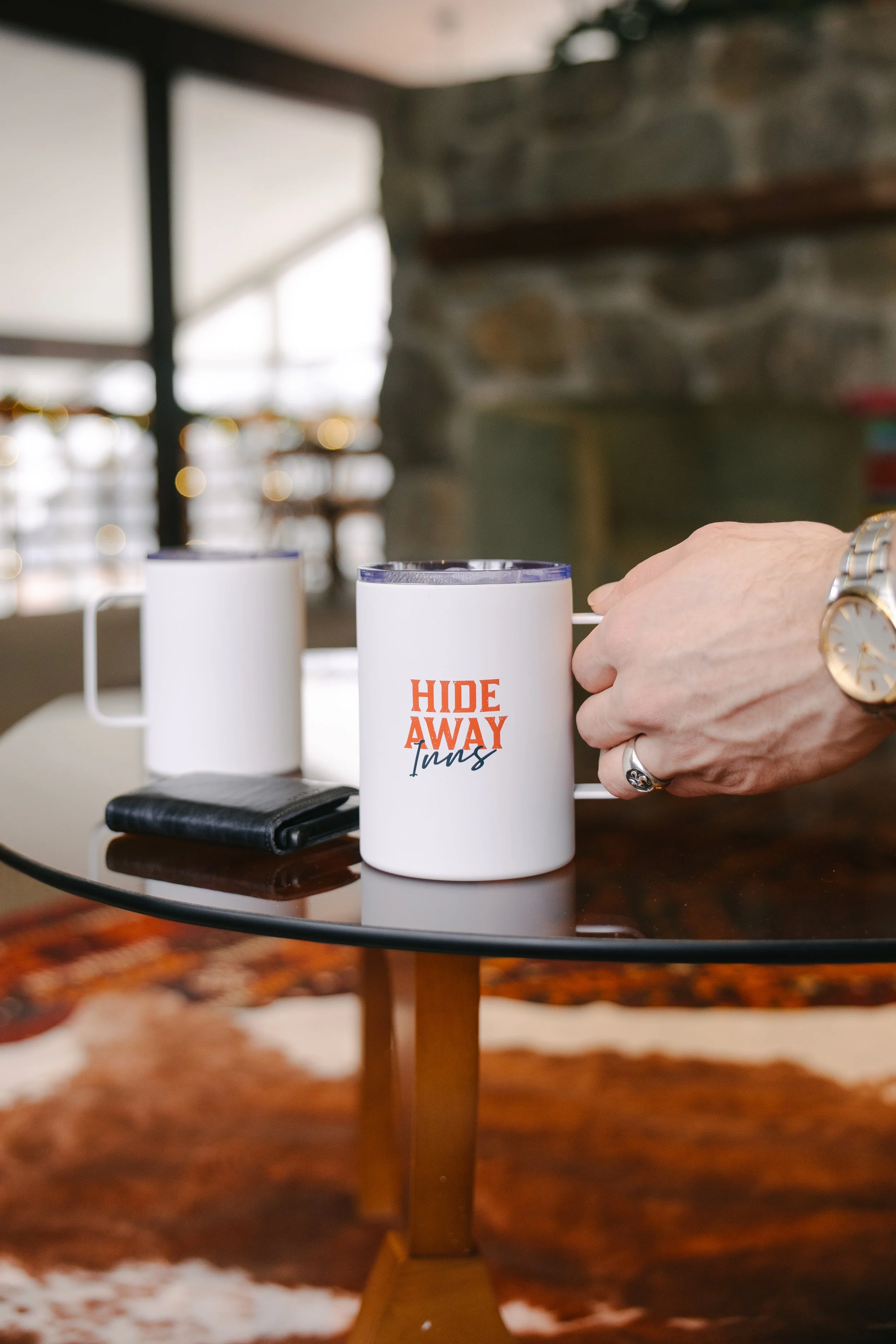 Two white coffee mugs on a wooden table, one with red and black text that reads 'HIDE AWAY Inn,' with a hand holding the mug. There is a black wallet and a smartphone next to the mugs, and a background of stone wall and large windows.