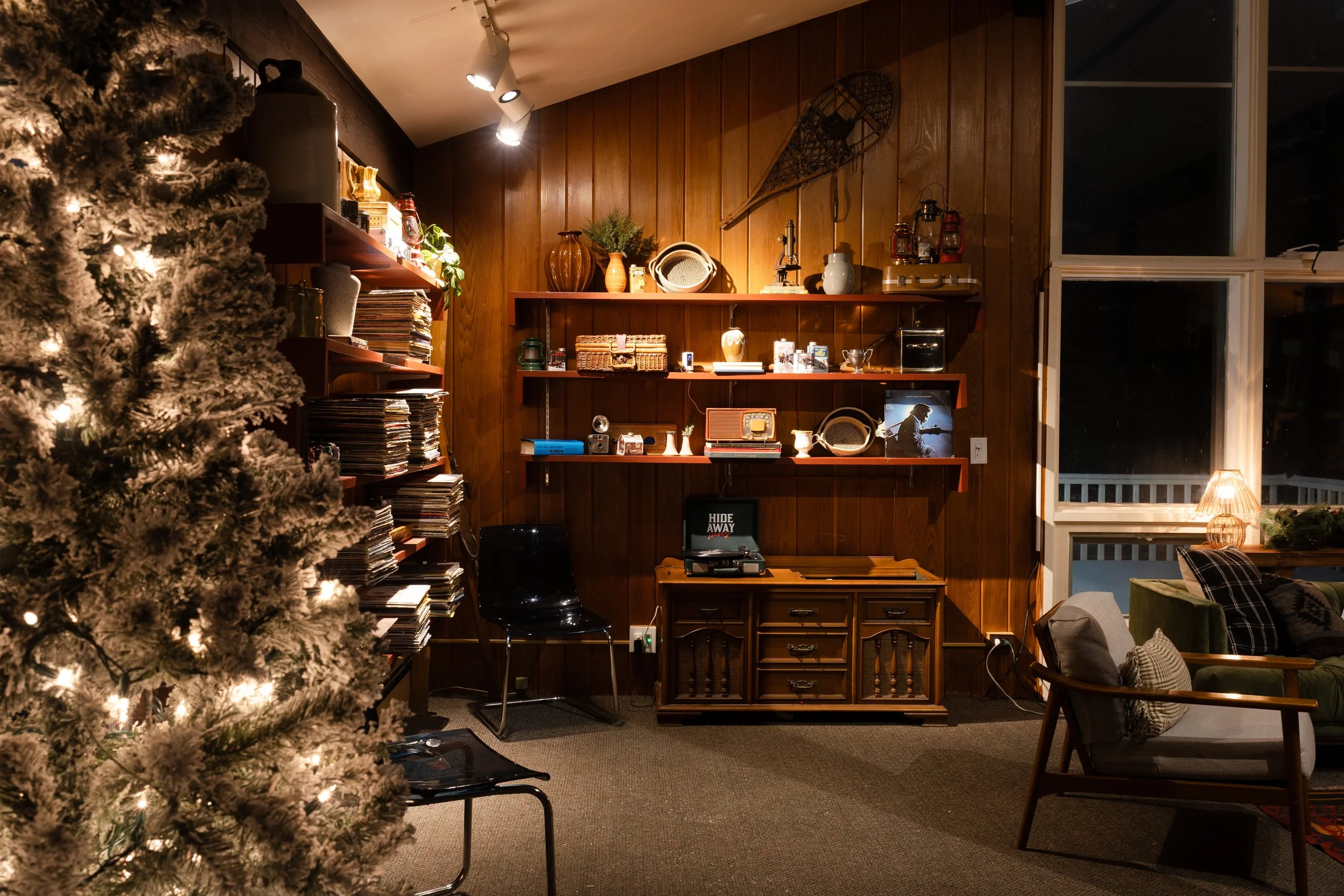 A cozy living room with a decorated Christmas tree on the left, wooden wall decor, shelves with vintage and collectibles, a window with night view, and warm lighting creating a festive atmosphere.