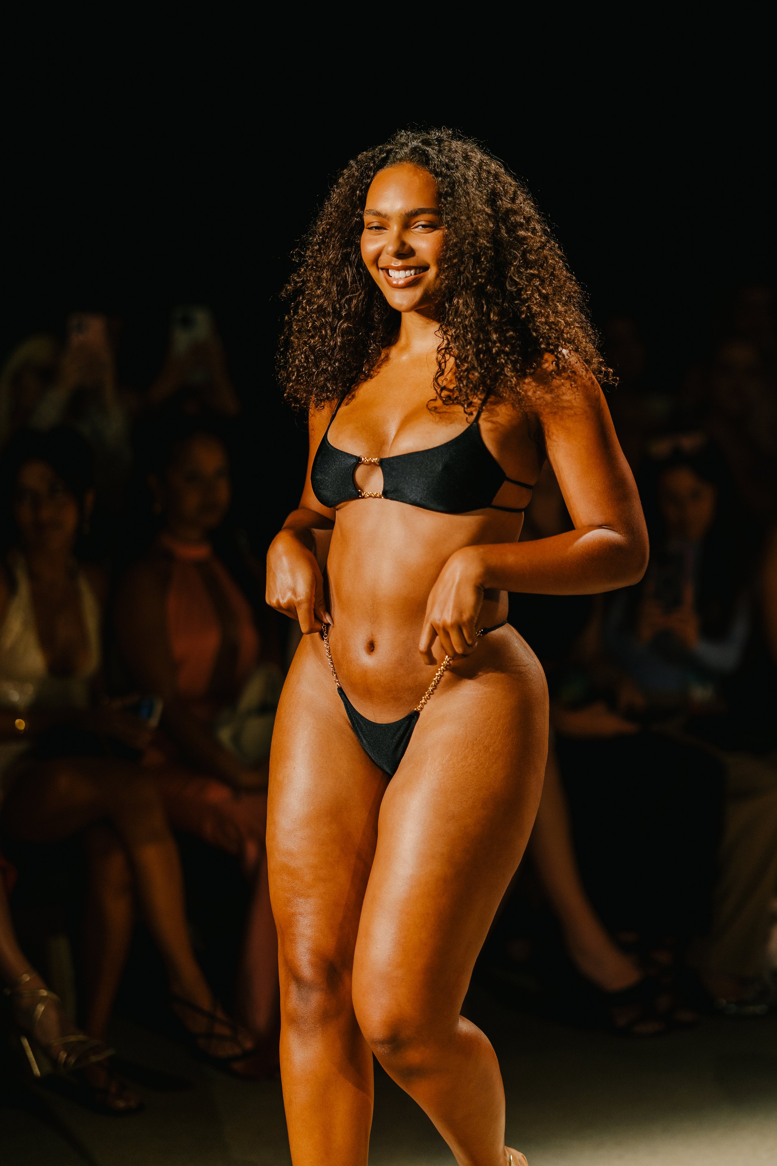 A woman walking on a runway in a black bikini with a chain detail, smiling as she models in front of an audience.