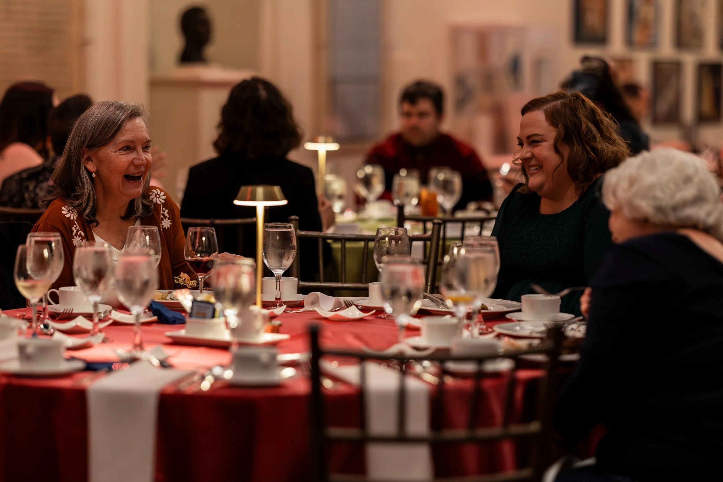People sitting at a table in a restaurant or banquet hall, laughing and enjoying a meal, with a blurred background of other diners and artwork on the walls.