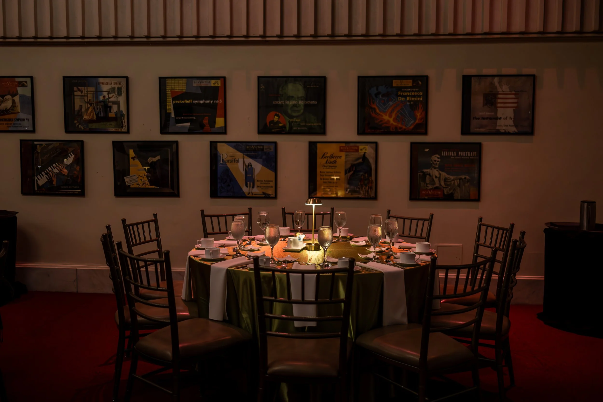 A restaurant dining area with a round table set with white tablecloth, wine glasses, water glasses, cups, and utensils. The table has a small lamp in the center. The background shows a white wall decorated with framed posters or artwork.