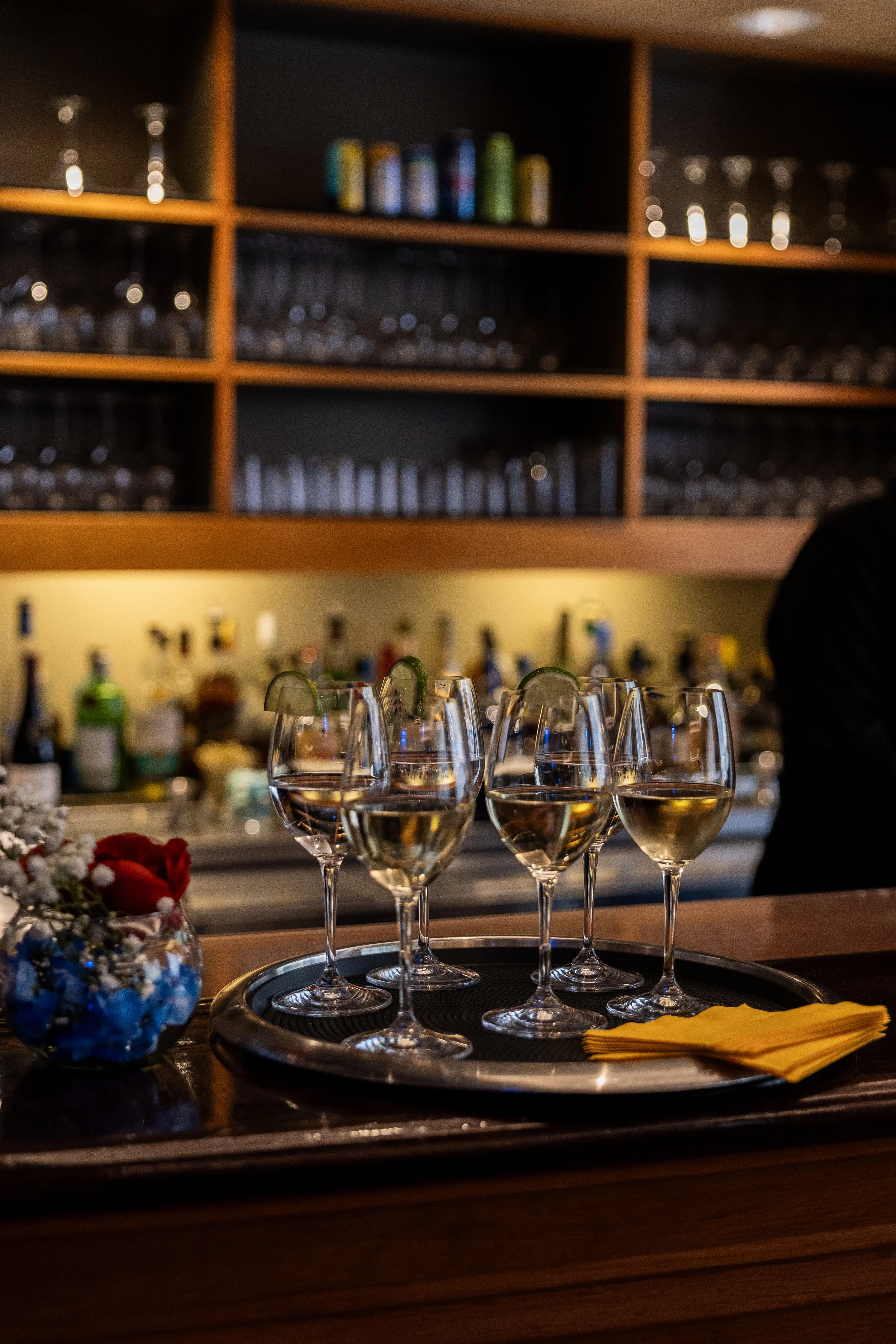 A bar or restaurant setting with several glasses of white wine on a tray, garnished with lime slices, and a yellow cloth napkin, with shelves of glassware and alcohol bottles in the background.