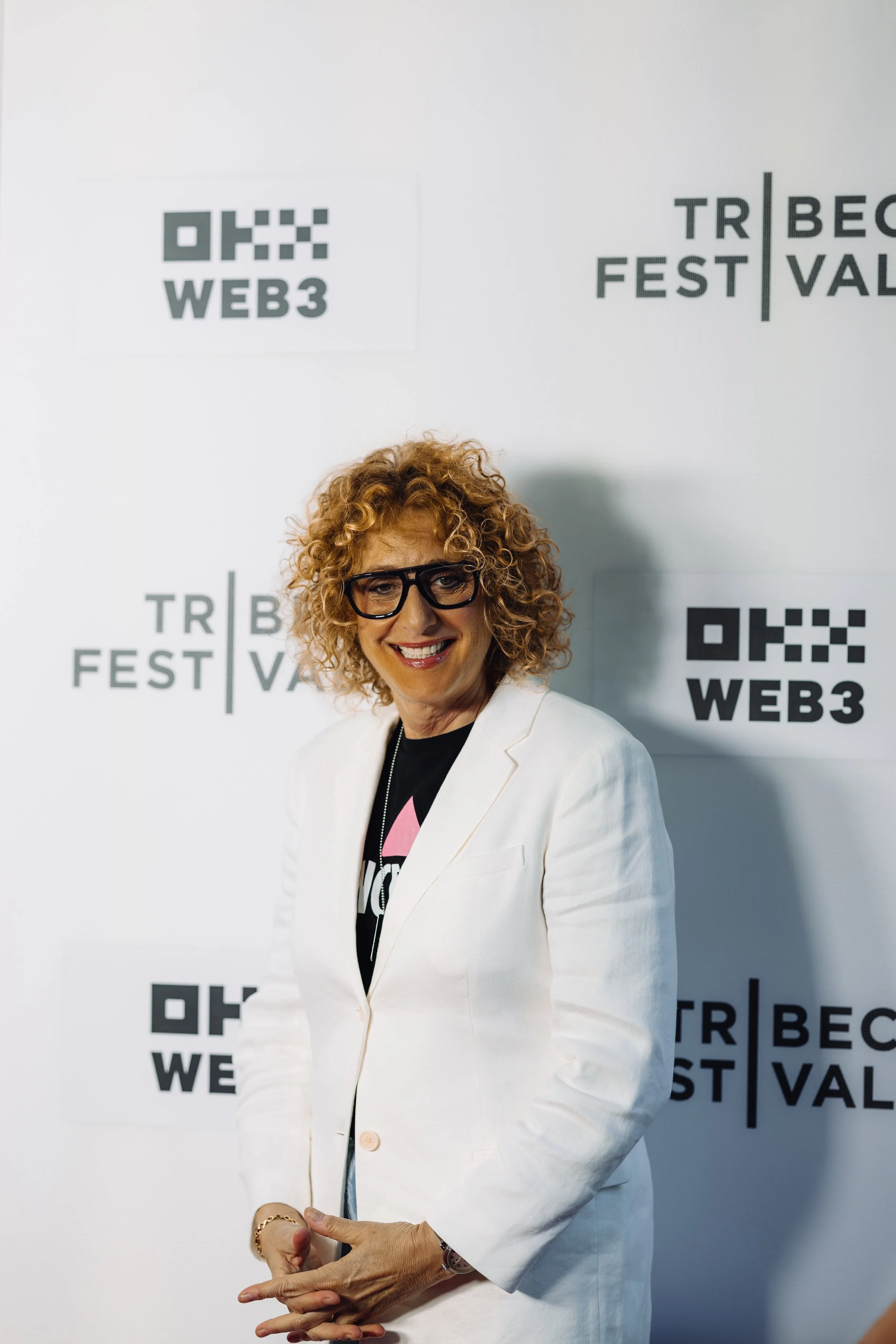 A woman with curly blonde hair and glasses smiling at the camera, wearing a white blazer over a black t-shirt with a pink graphic. She is standing in front of a white backdrop with black logos and text for TRIBECA FILM FESTIVAL and OKX WEB3.