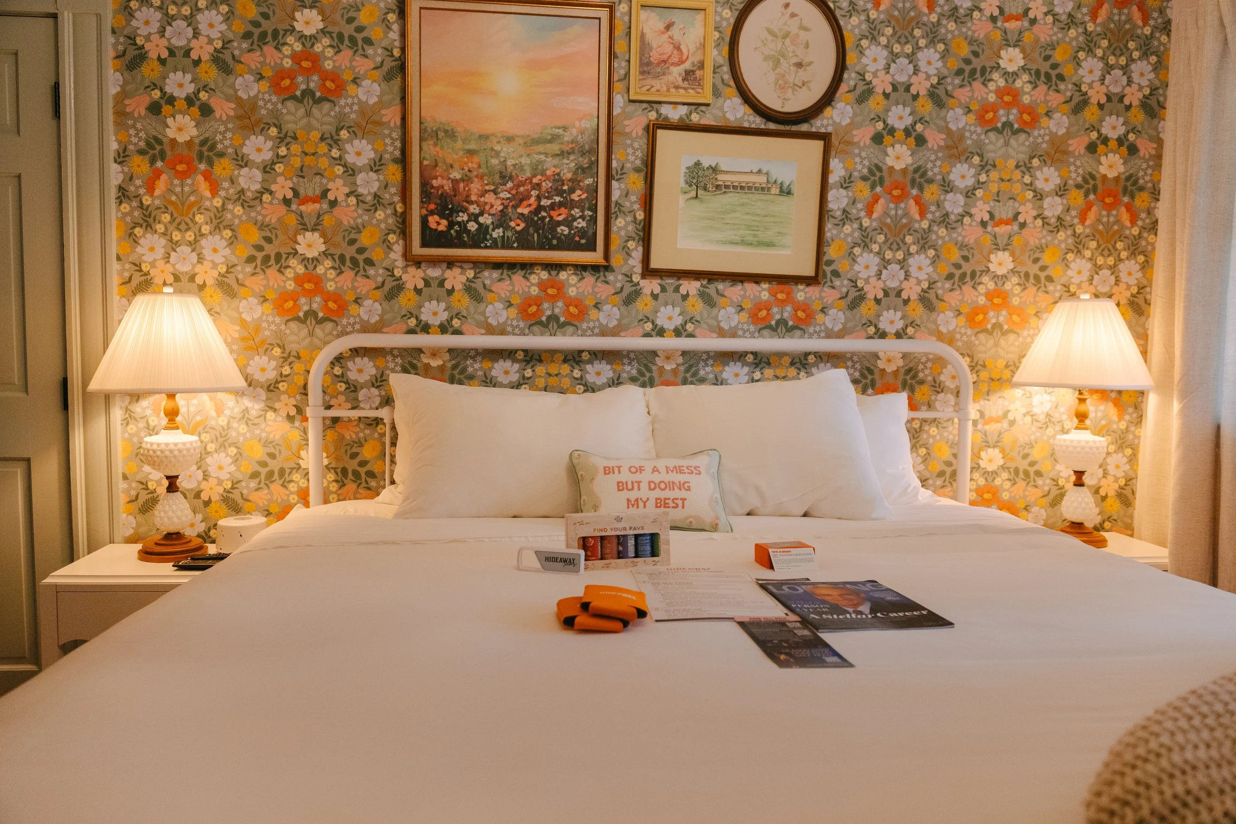 Cozy bedroom with floral wallpaper, two white bedside lamps, a white bed with a pillow that reads 'Bit of a mess but doing my best', and several items on the bed including magazines, brochures, and a small box.