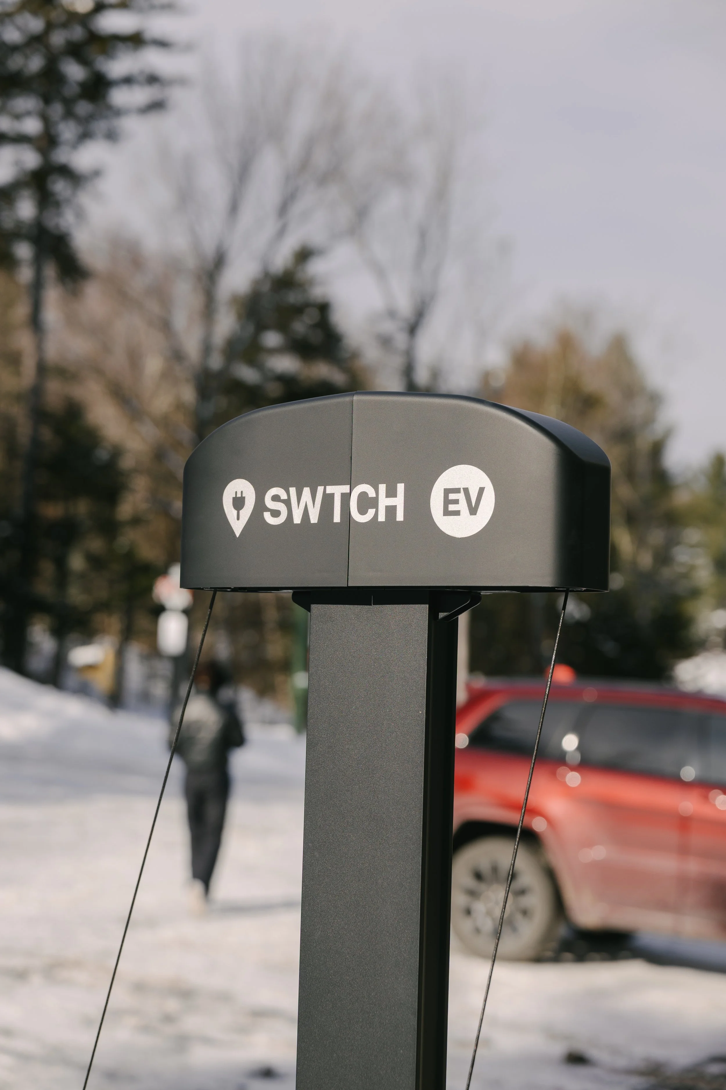 An electric vehicle charging station with a sign that reads 'SWITCH EV' in an outdoor snowy setting. In the background, a person and a red vehicle are visible.