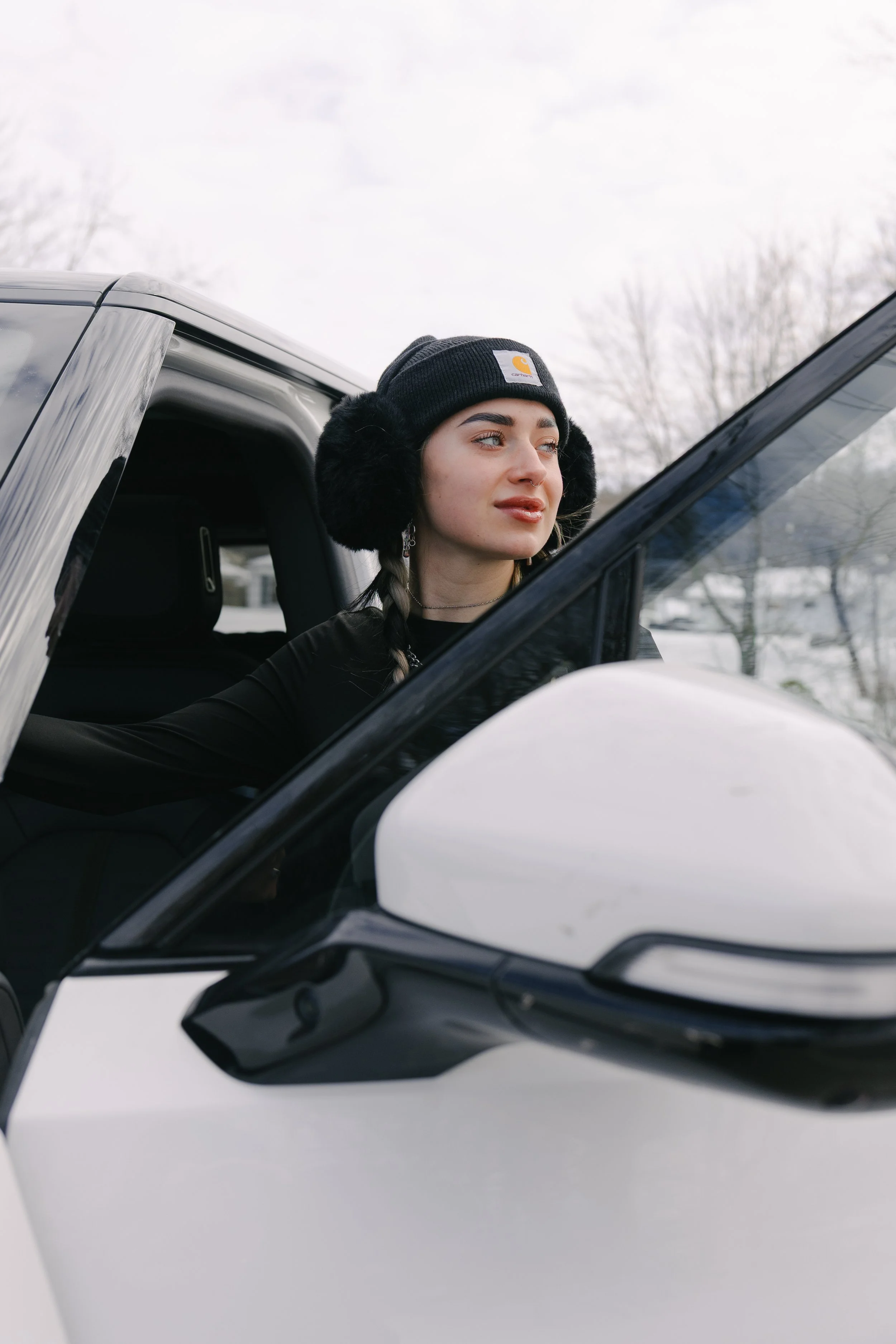 A young woman wearing a black beanie with a Carhartt logo, black earmuffs, and a black shirt, looking out of a car window on a snowy day.