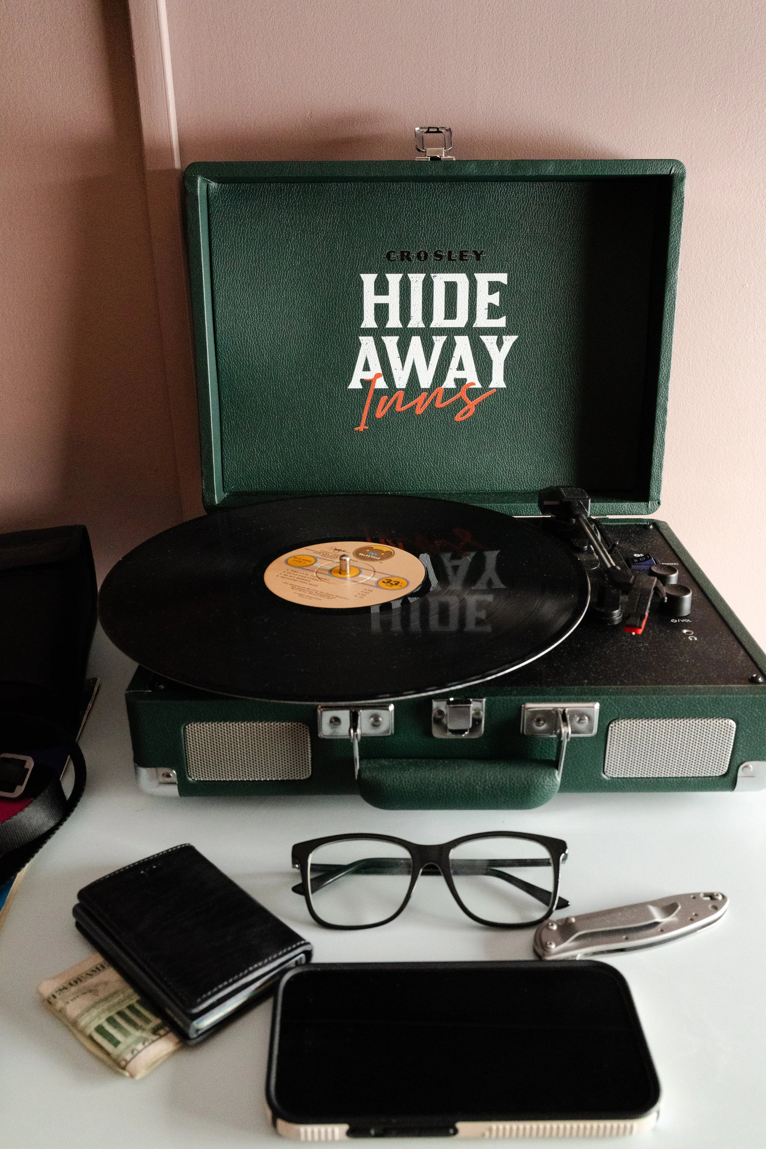 A vintage green portable record player playing a vinyl record, with sunglasses, a wallet with cash, a smartphone, a knife, and a black bag nearby on a white surface.