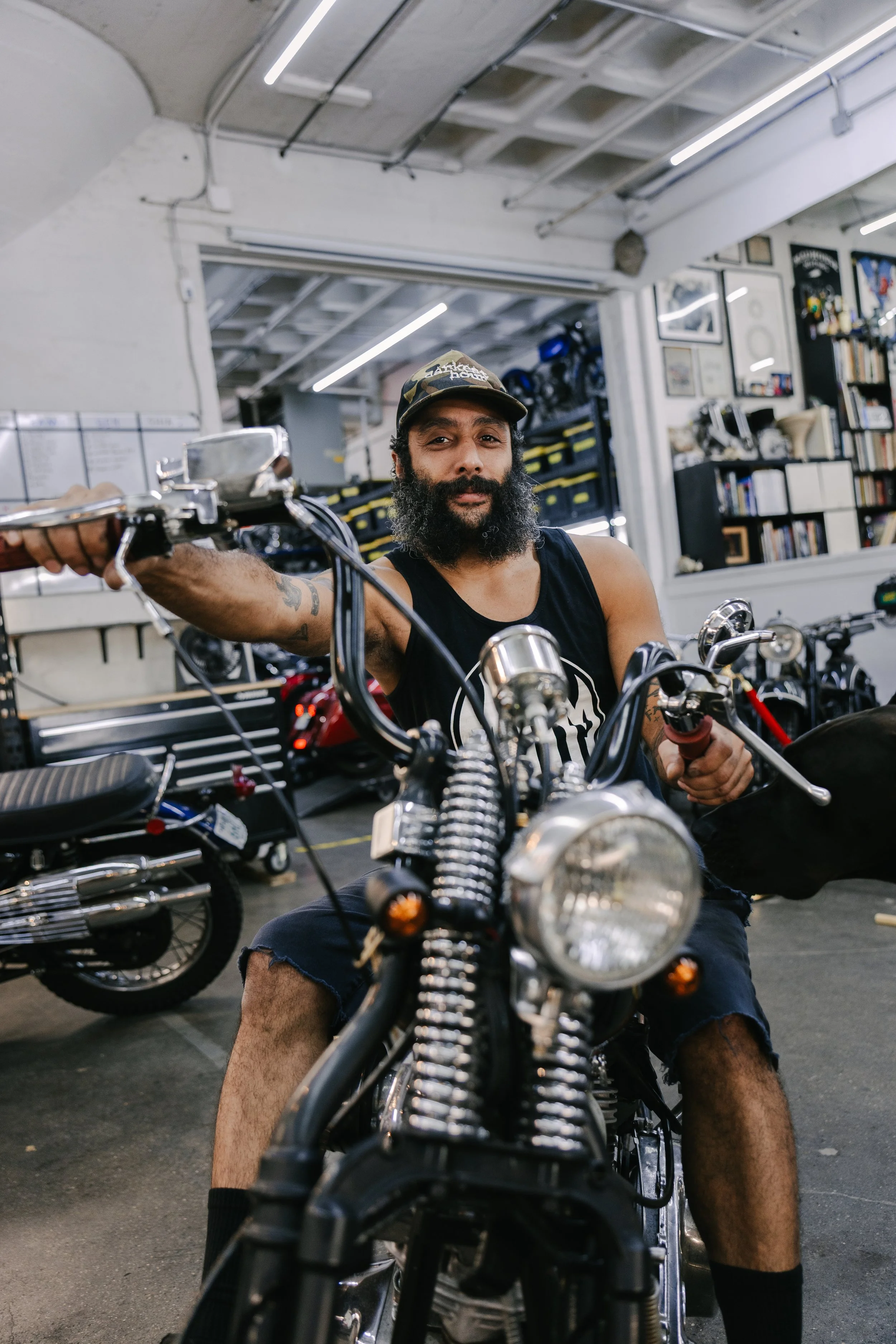 Man sitting on a motorcycle in a workshop surrounded by tools, shelves, and motorcycle parts.