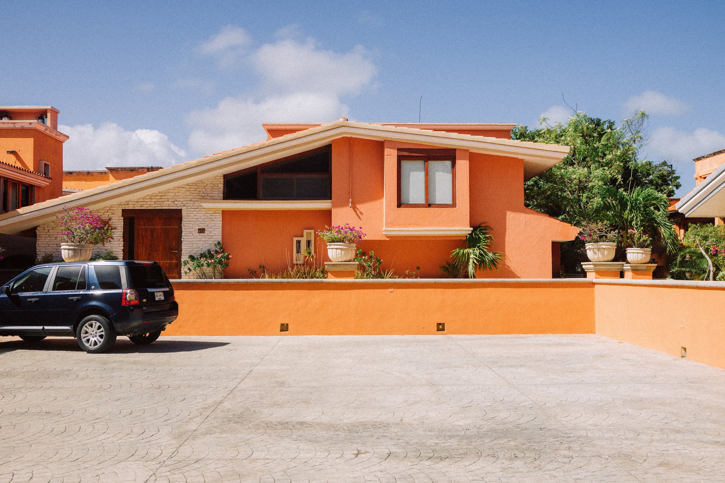 A modern orange house with a sloped roof, surrounded by plants, with a black car parked in front on a paved area.