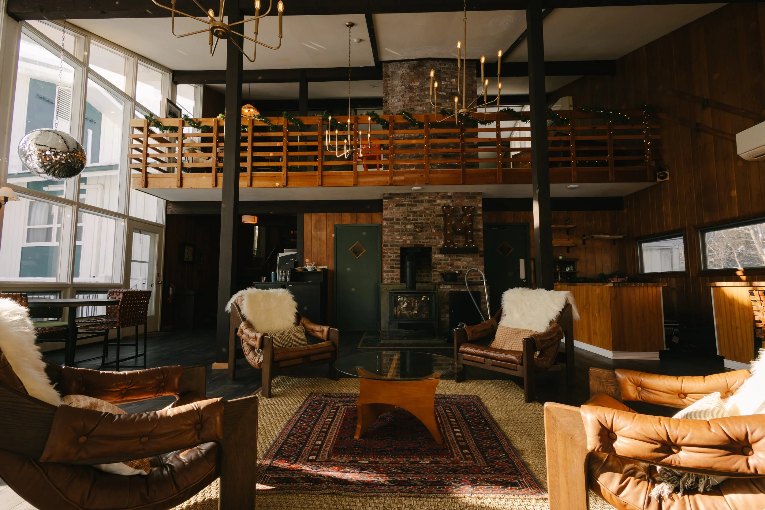 Cozy interior of a living room with leather armchairs, a glass coffee table, a brick fireplace, wooden walls, large windows, and a second-story balcony decorated with green garland and string lights.