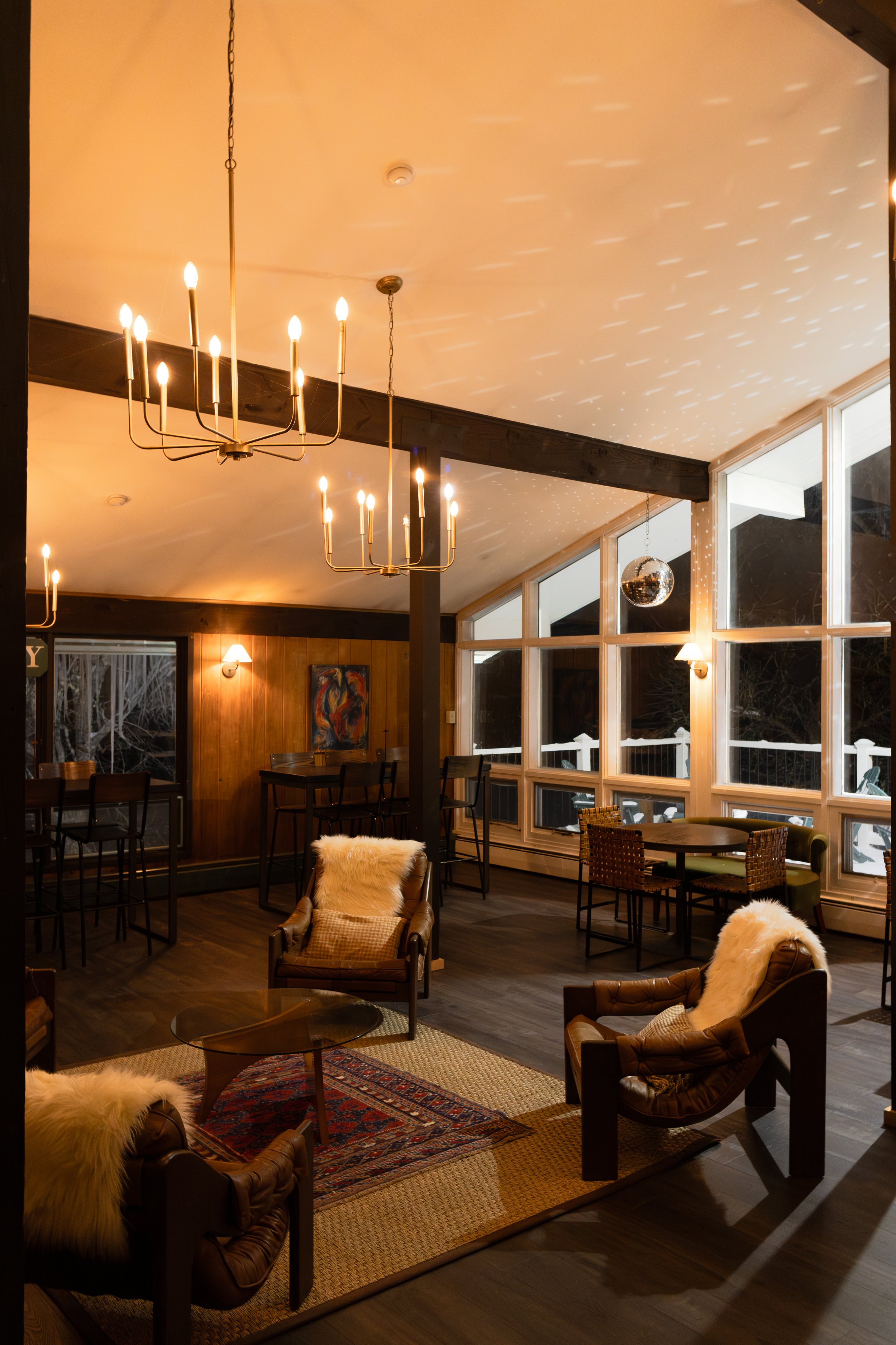 Cozy living room with leather armchairs, fluffy throws, a rug, large windows, and warm lighting including chandeliers and wall sconces.
