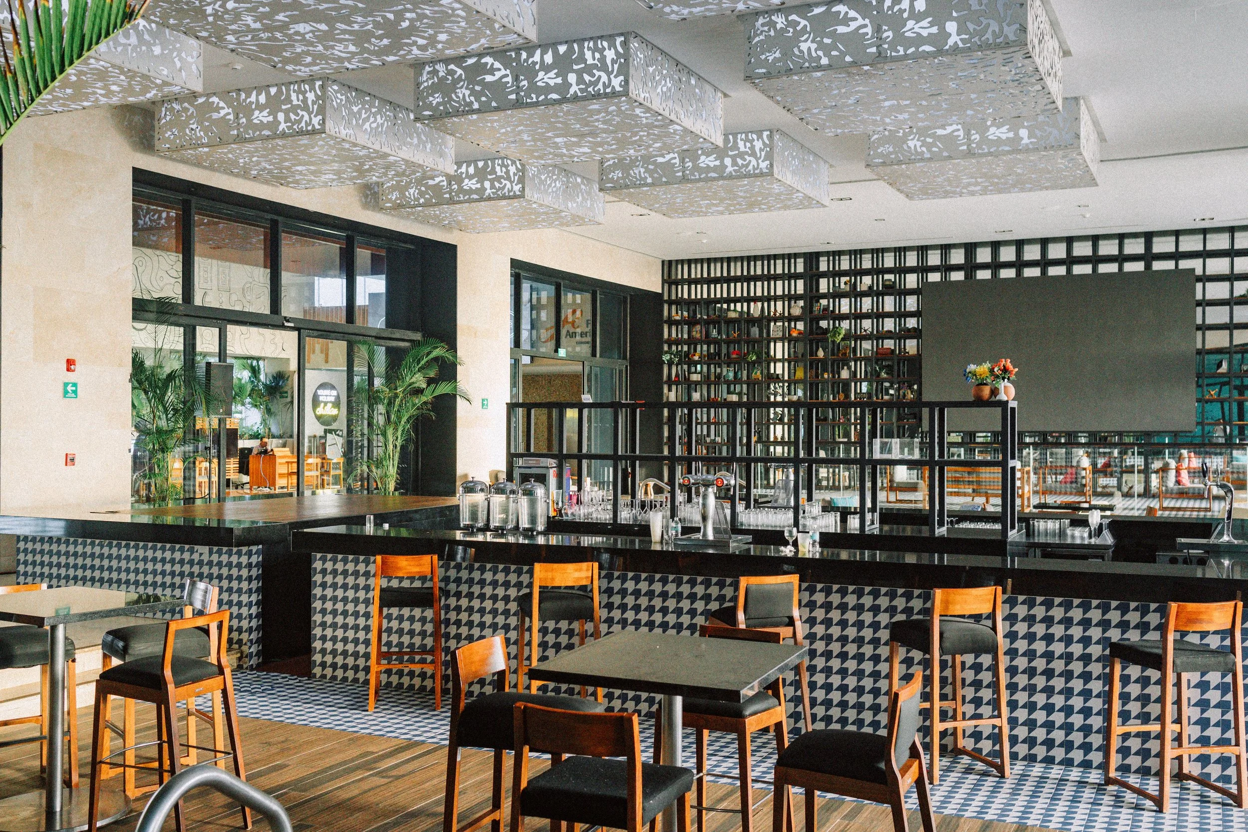 Interior of a modern restaurant with wooden chairs, black tables, decorative ceiling panels, and a black lattice wall with shelves and potted plants.