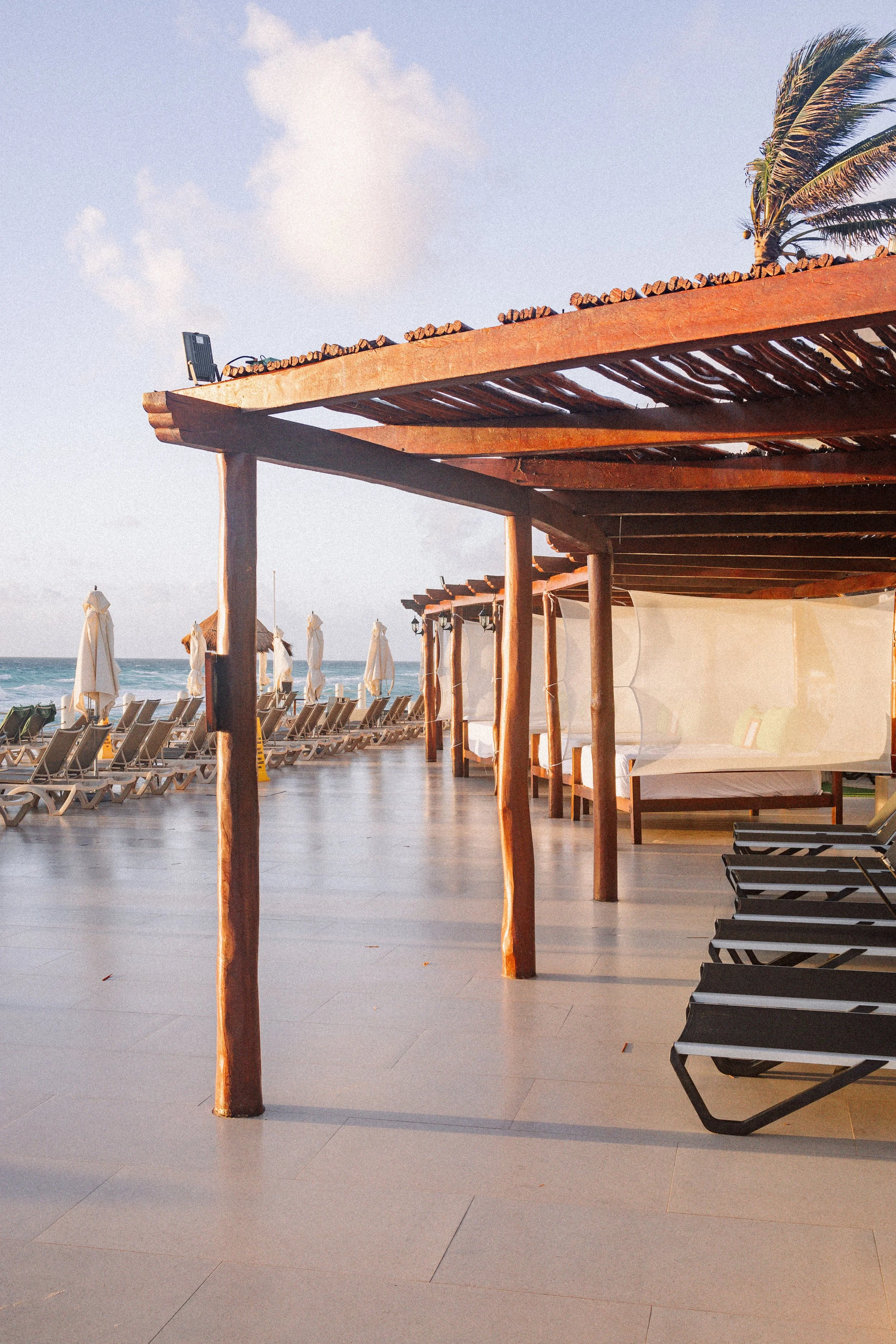 Beachside lounge chairs under a wooden cabana with umbrellas and a palm tree in the background, near the ocean.