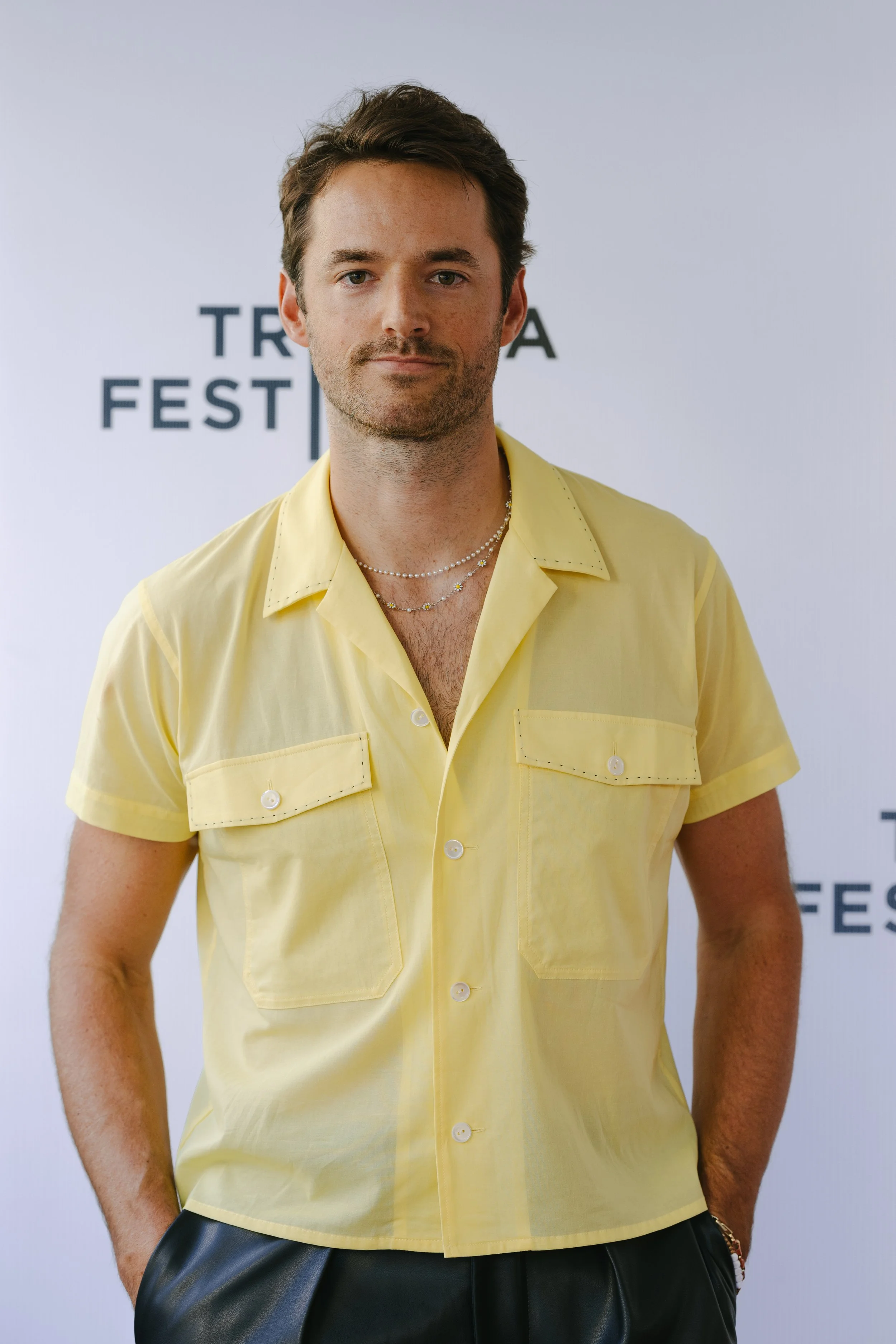 A man with brown hair and light stubble wearing a yellow short-sleeve shirt and layered necklaces, standing in front of a white backdrop with black text.