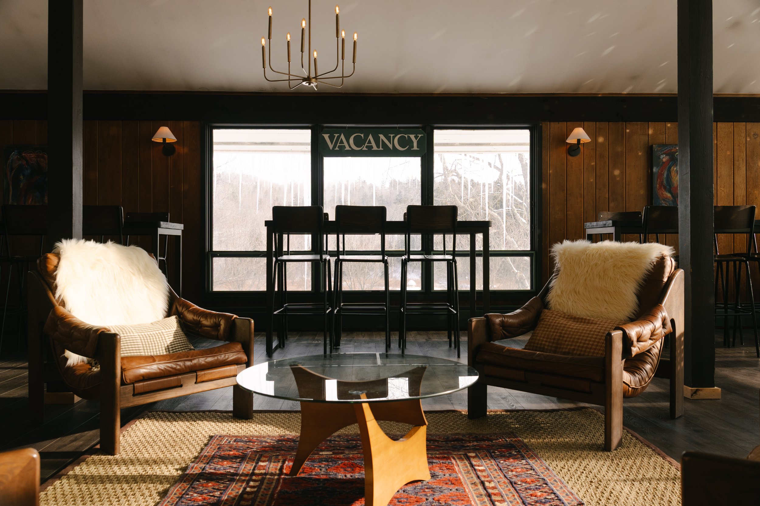 Indoor seating area with leather armchairs decorated with sheepskin throws and pillows, a glass-top coffee table, and tall bar stools in front of large windows with a three-pane design and a "VACANCY" sign, warm wooden wall paneling, and subdued ligh