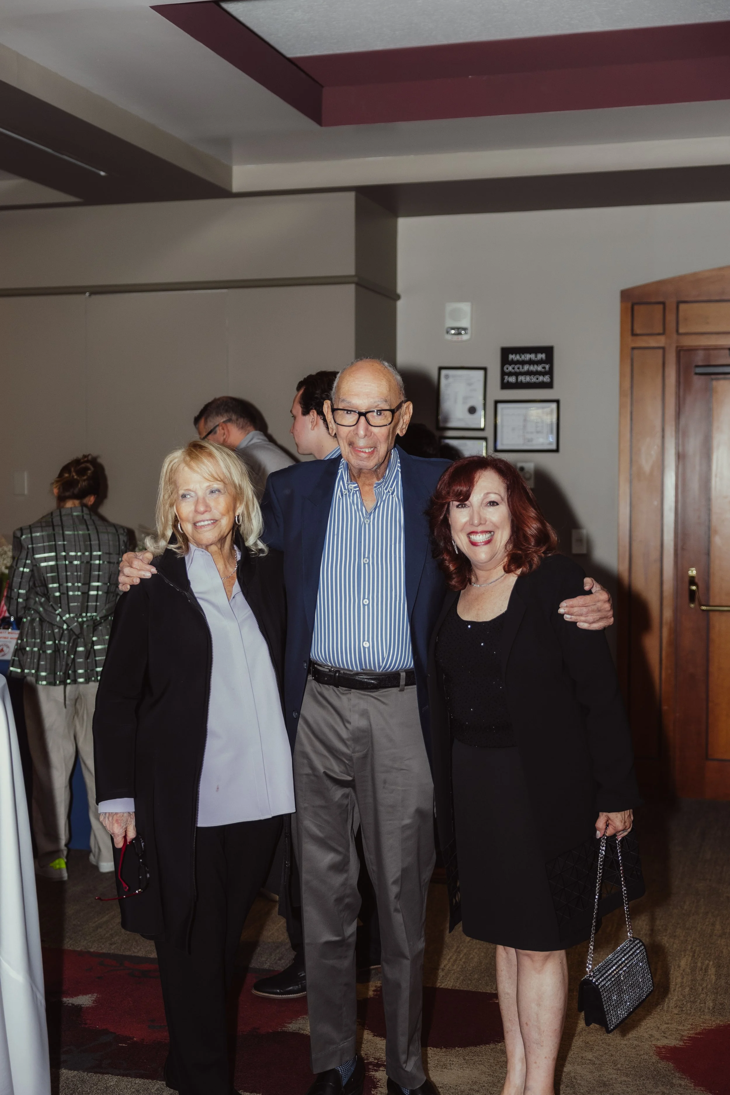 Three smiling people standing together at an indoor event, with other guests in the background. The man in the center is older with glasses, wearing a blue jacket and striped shirt. The woman on the left has light blonde hair and is dressed in black 