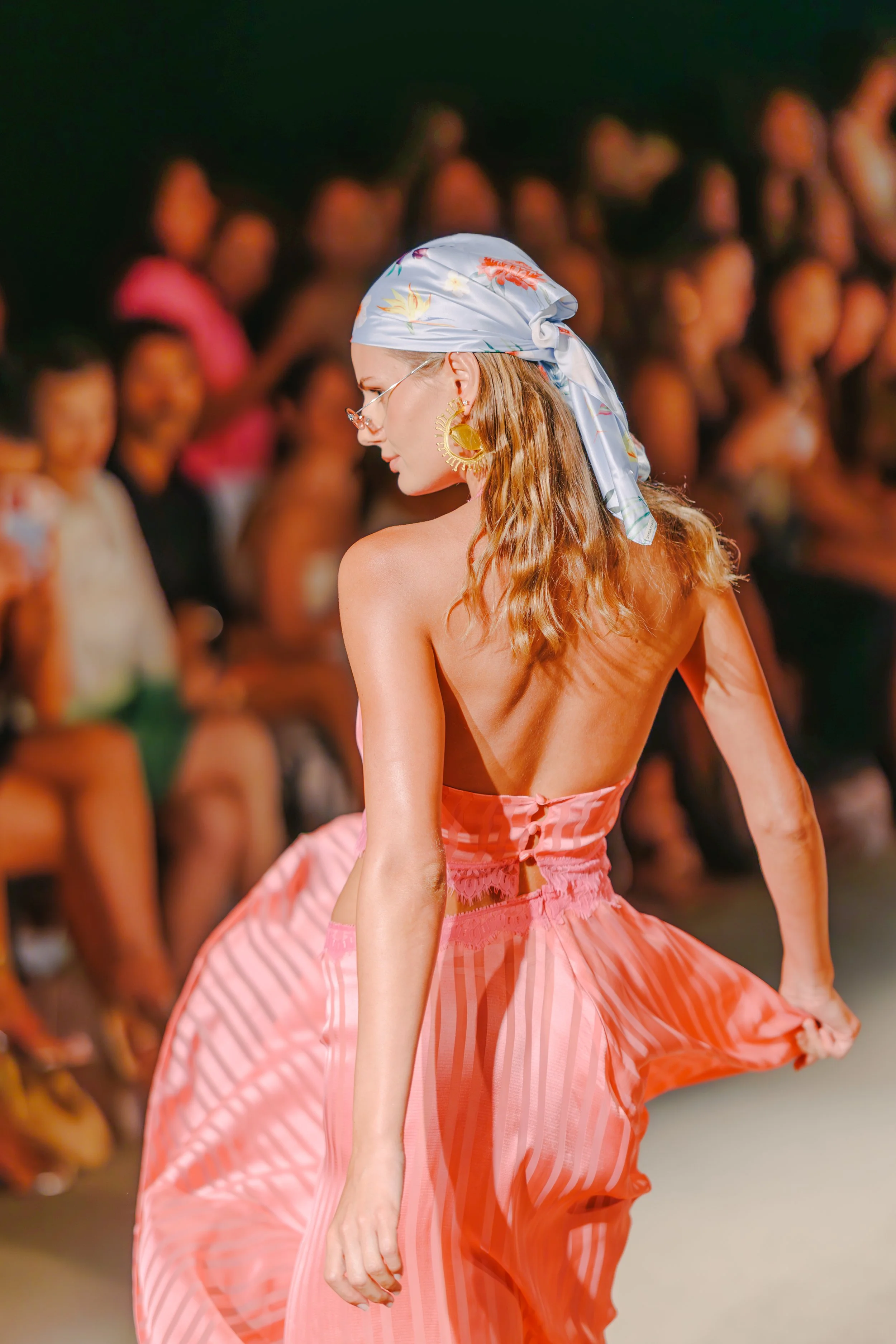 A woman with wavy hair wearing a satin headscarf, sunglasses, and large earrings, walking on a runway in a pink, striped dress with an open back, while an audience watches in the background.
