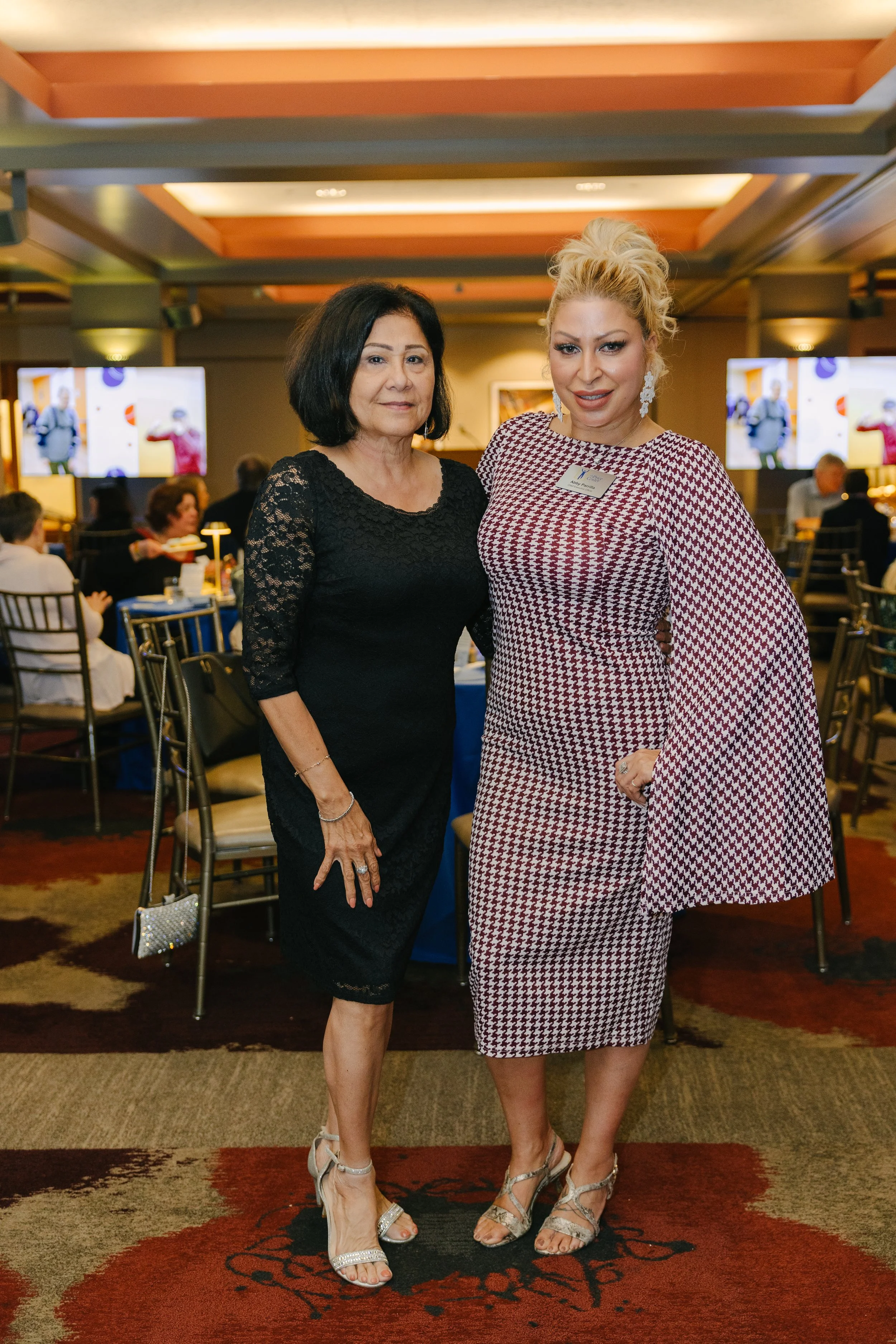 Two women standing together at a formal event, smiling and posing for the photo. The woman on the left has short dark hair and is wearing a black lace dress, while the woman on the right has blonde hair styled up and is dressed in a red and white pat