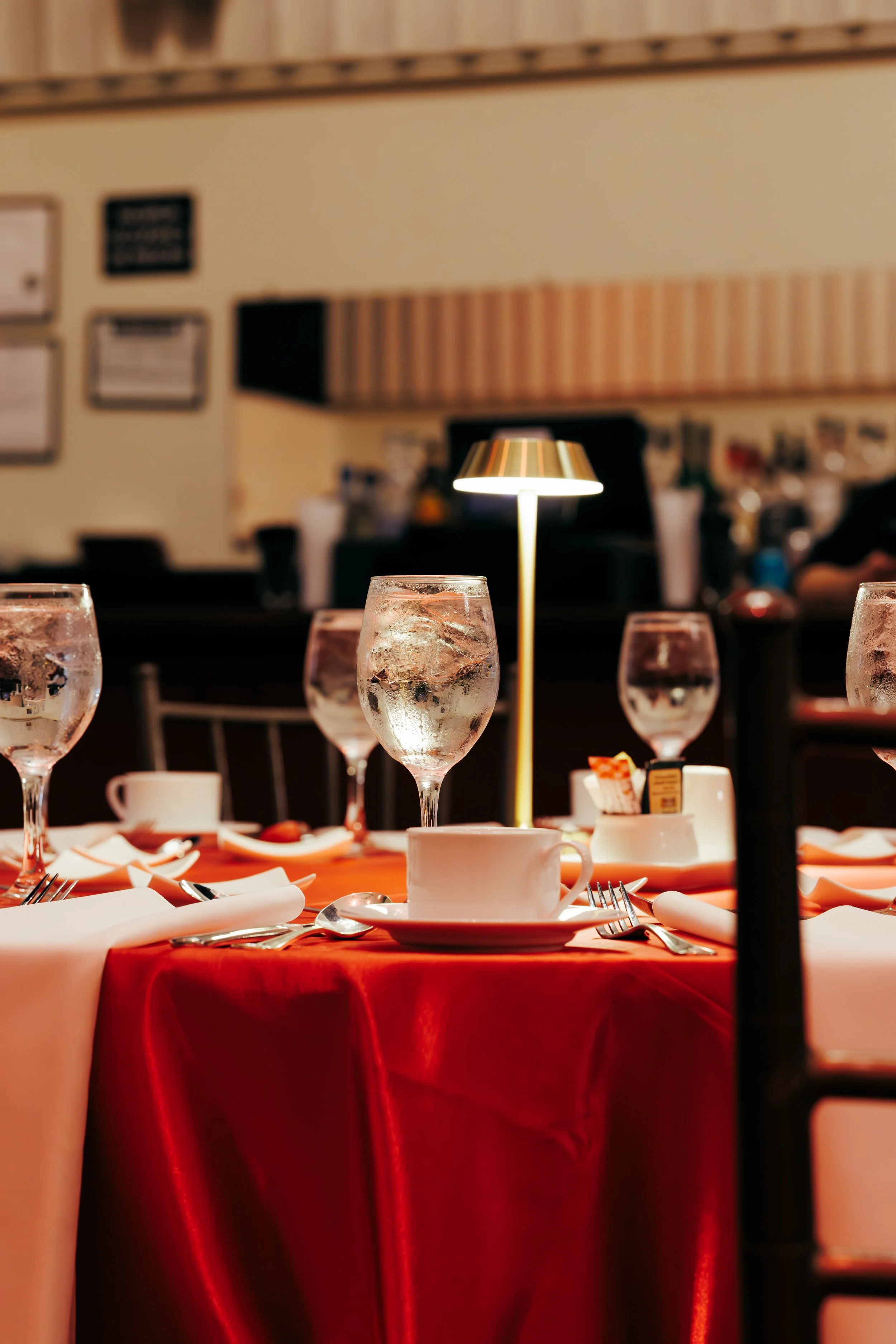 Table set for a meal with water glasses, white cups, napkins, and silverware, under a table lamp, in a restaurant with framed pictures on the wall.