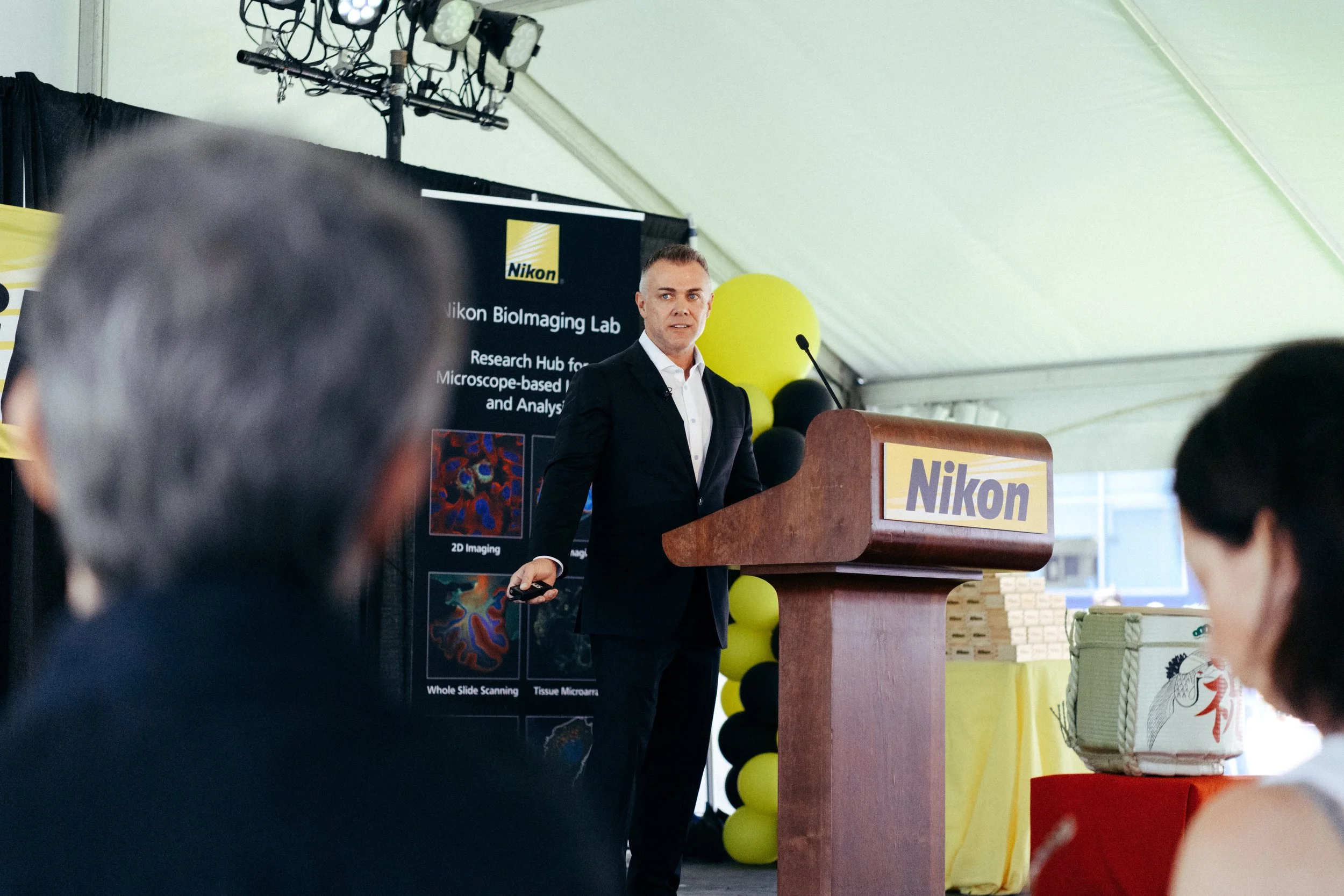Man giving a presentation at a Nikon event with a podium and a large display behind him that says 'Nikon Biolmaging Lab'. The audience is visible in the foreground, and there are yellow and black balloons behind the speaker.