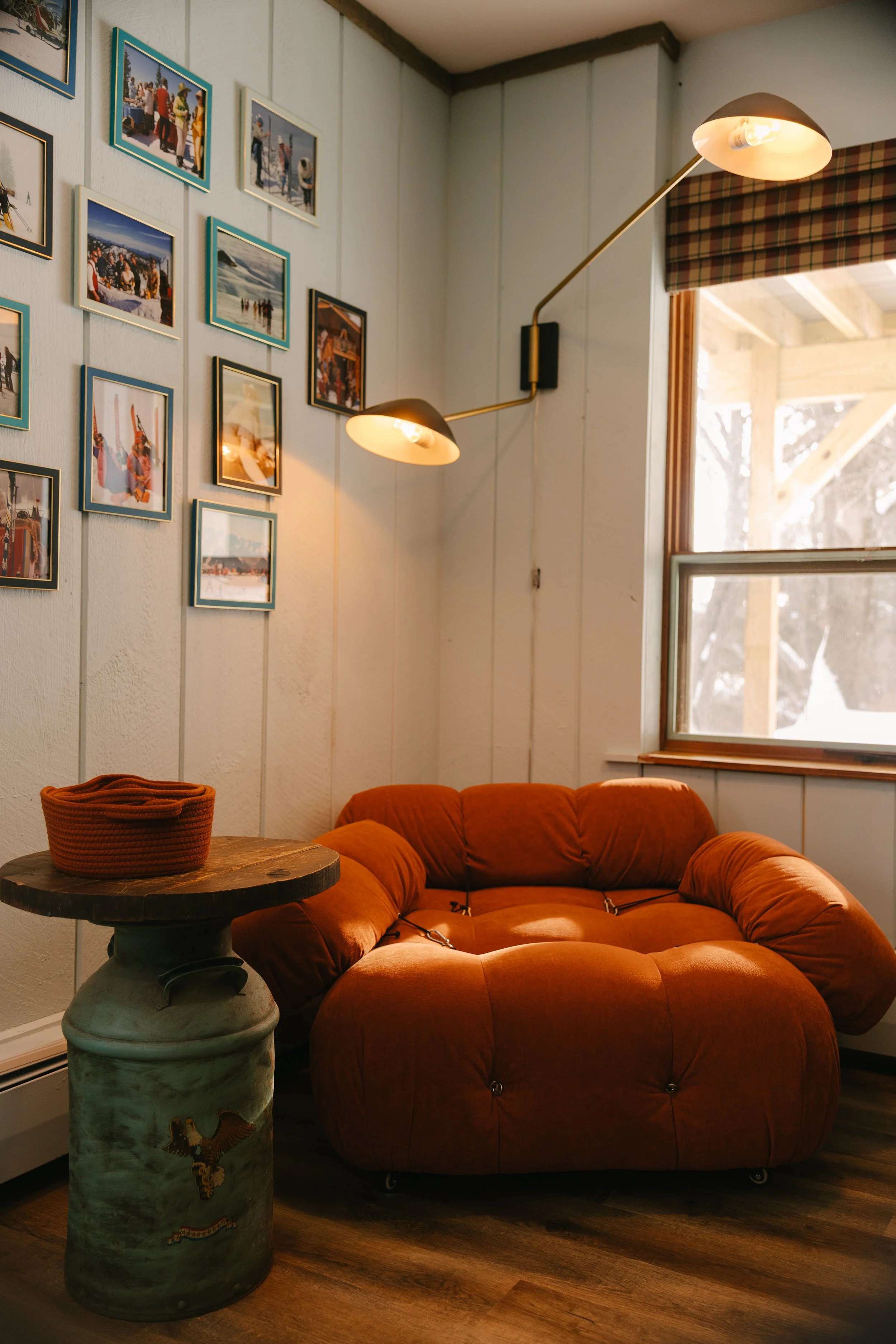 A cozy corner of a living room with a rust-colored, plush, round sofa near a window. A wall with several framed photographs and postcards is illuminated by a corner wall-mounted lamp. A wooden table with a ceramic base, decorated with a small embroid