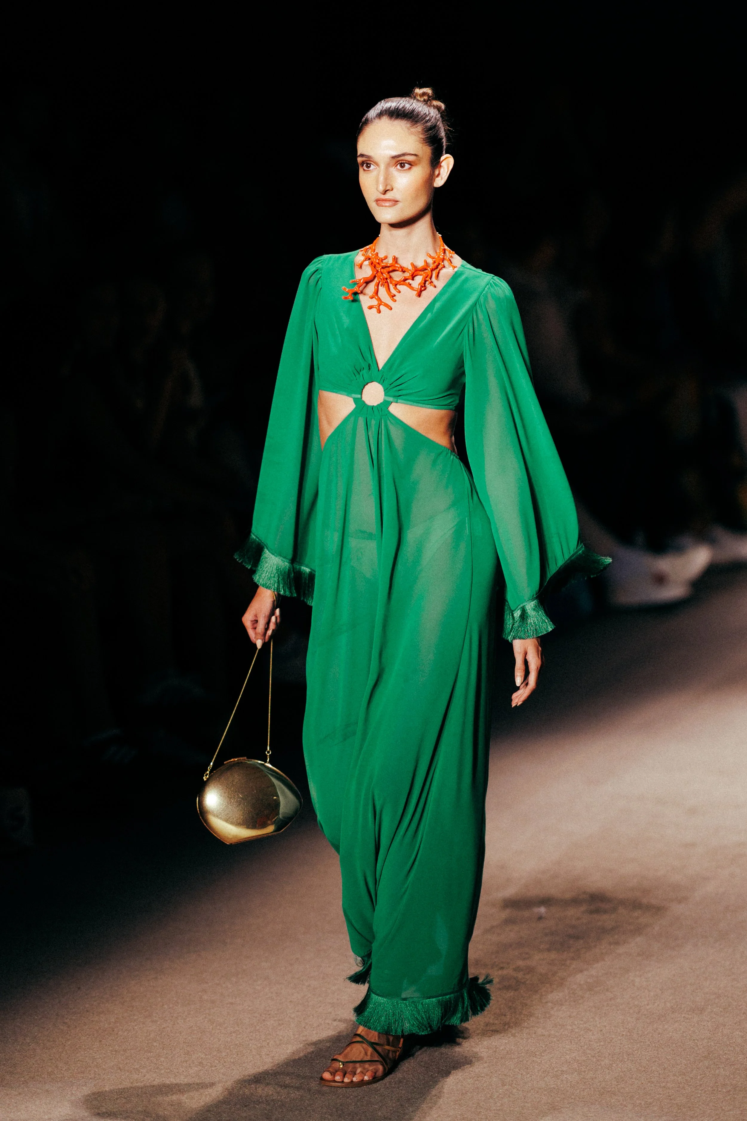 A female model walking on a runway wearing a green dress with cutouts and a circle detail at the waist, paired with a coral necklace, black sandals, and holding a small round golden handbag.