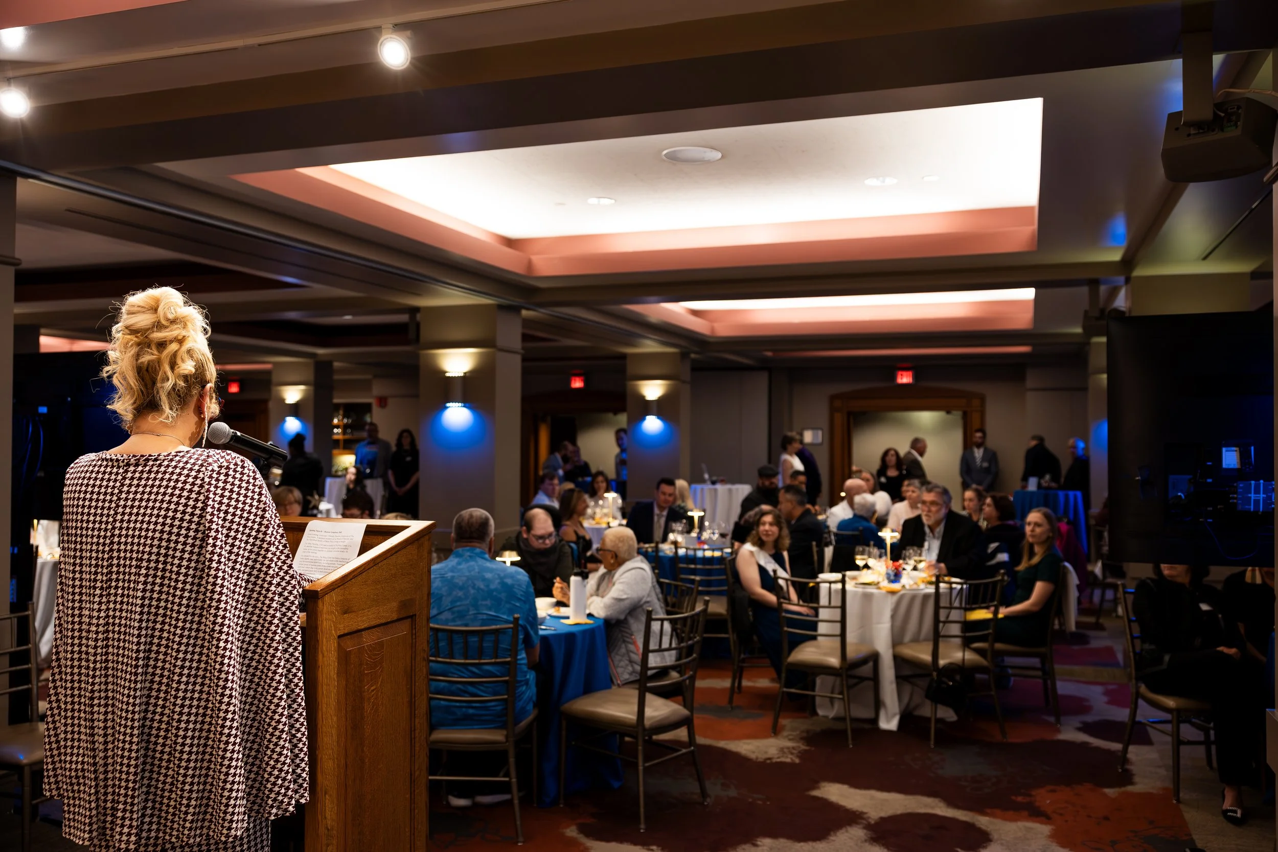 Woman with curly blonde hair speaking at a podium in front of a large audience at an indoor event with tables and guests
