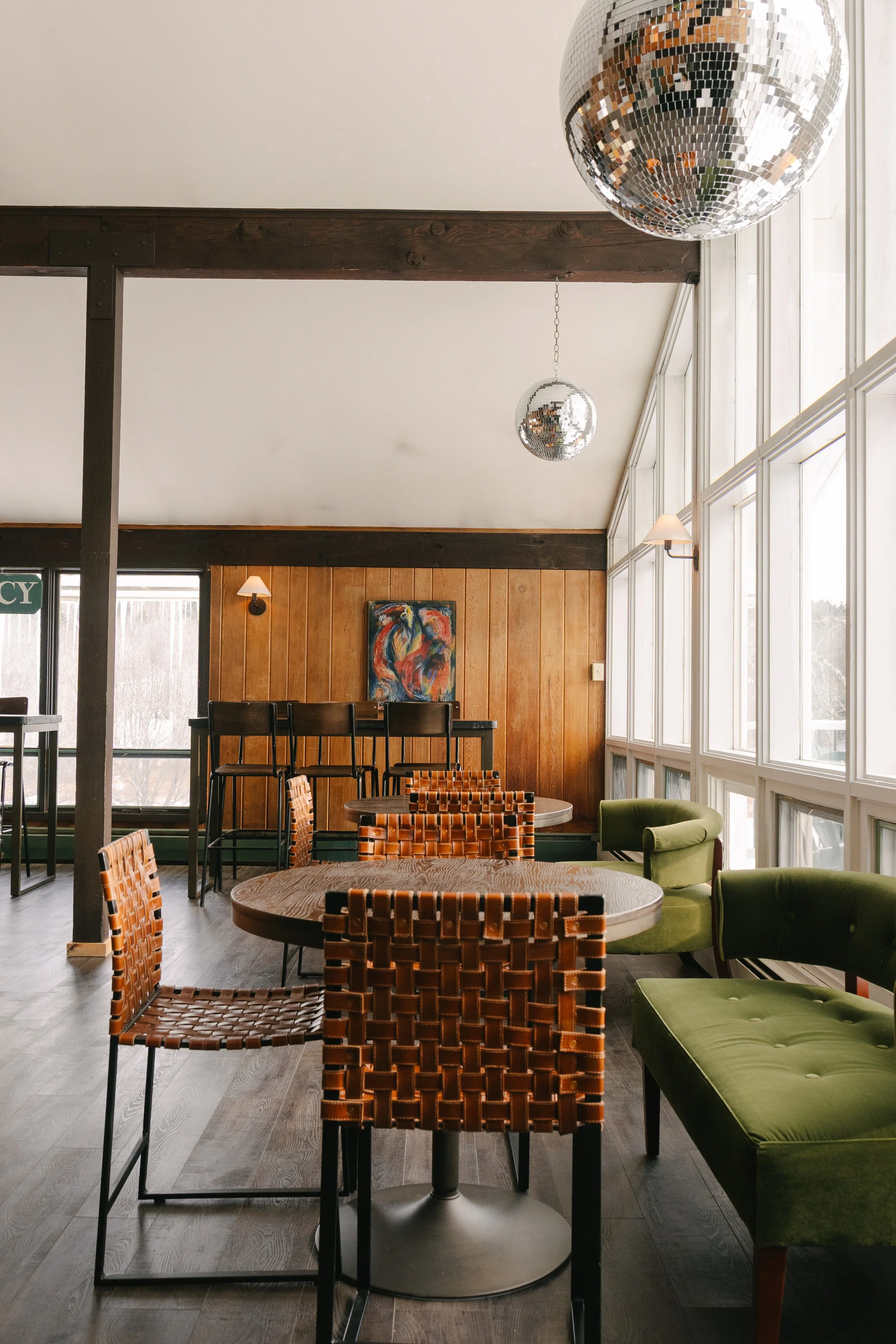 Indoor seating area with wooden and green upholstered chairs, wooden tables, wood-paneled walls, large windows, wall art, and disco balls hanging from the ceiling.