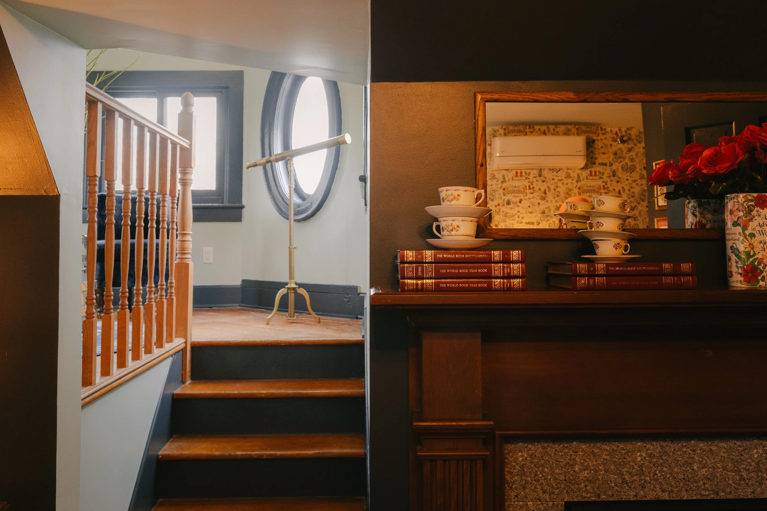 Interior of a cozy home with wooden stairs, a mirror, a window with round black trim, and a bar or counter with teacups, bowls, a stack of books, and a potted plant.