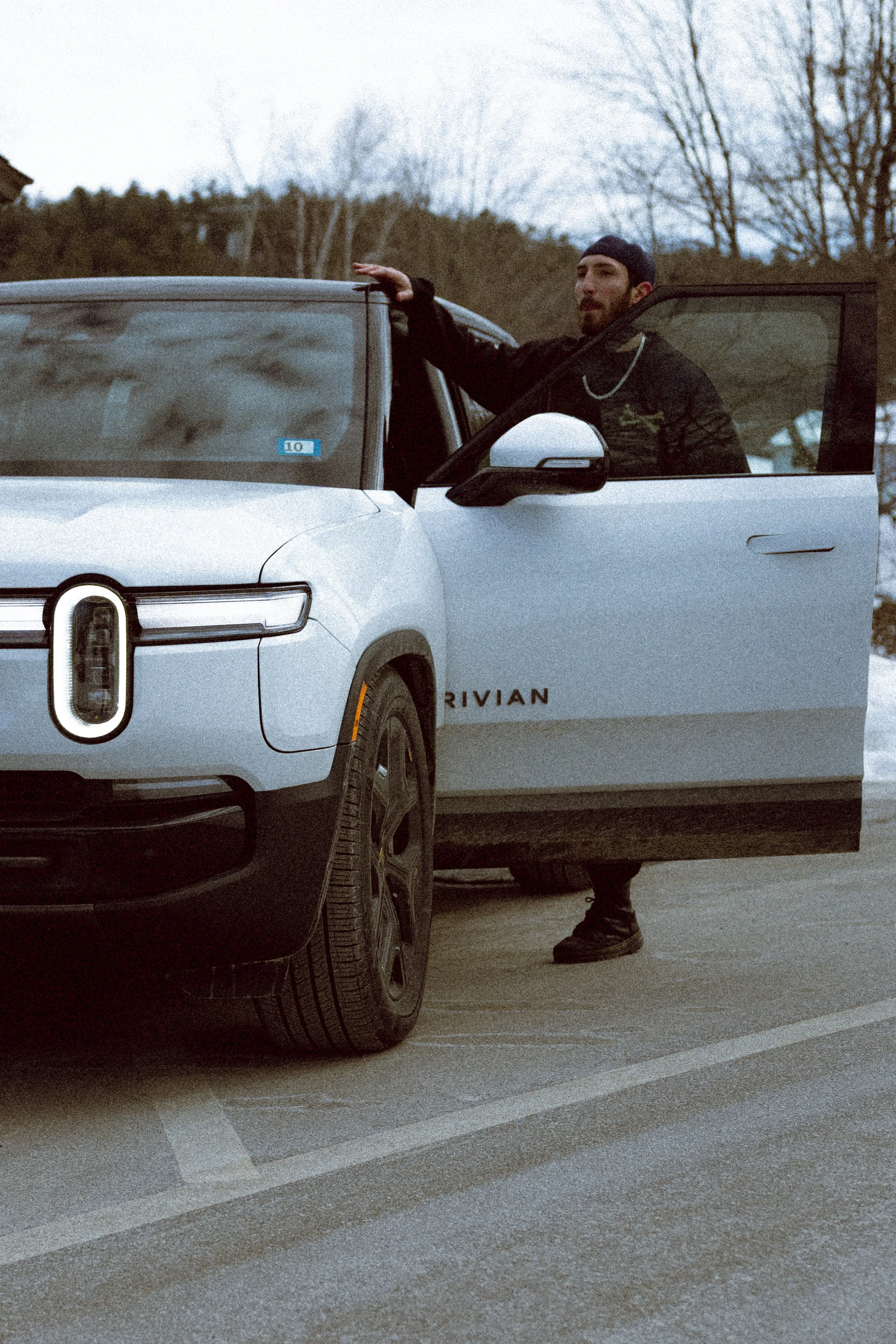 A man standing outside a white Rivian electric vehicle with one of the doors open, parked in a lot with trees in the background.