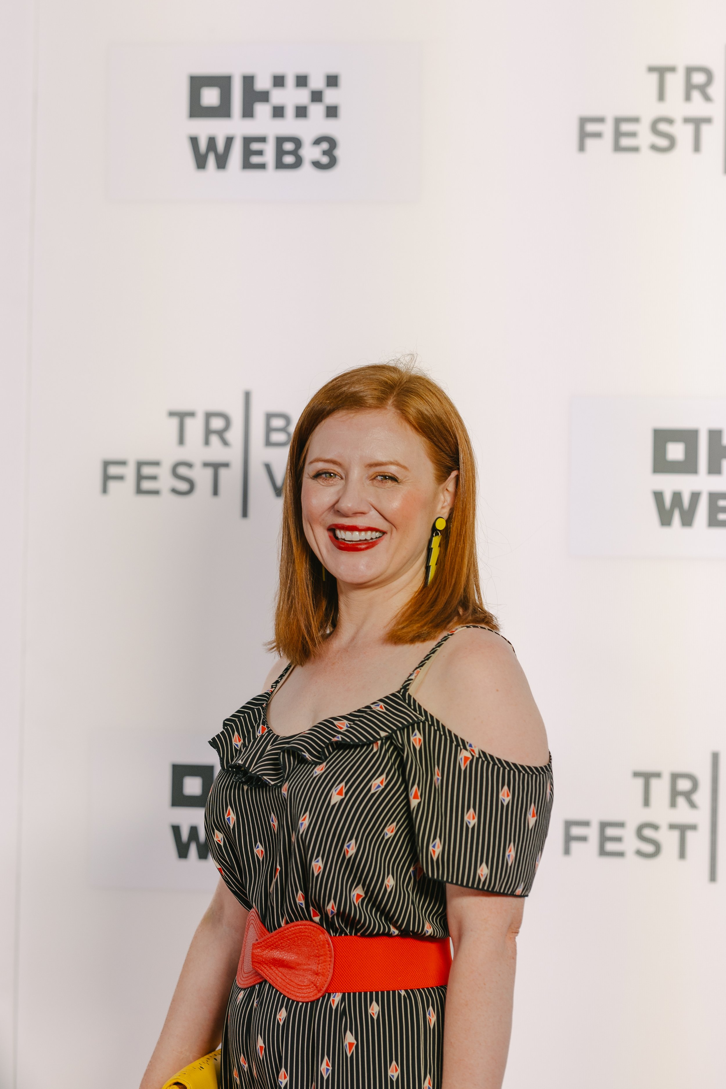 A woman with red hair smiling at the camera at an event, standing in front of a backdrop with logos for web and festival.