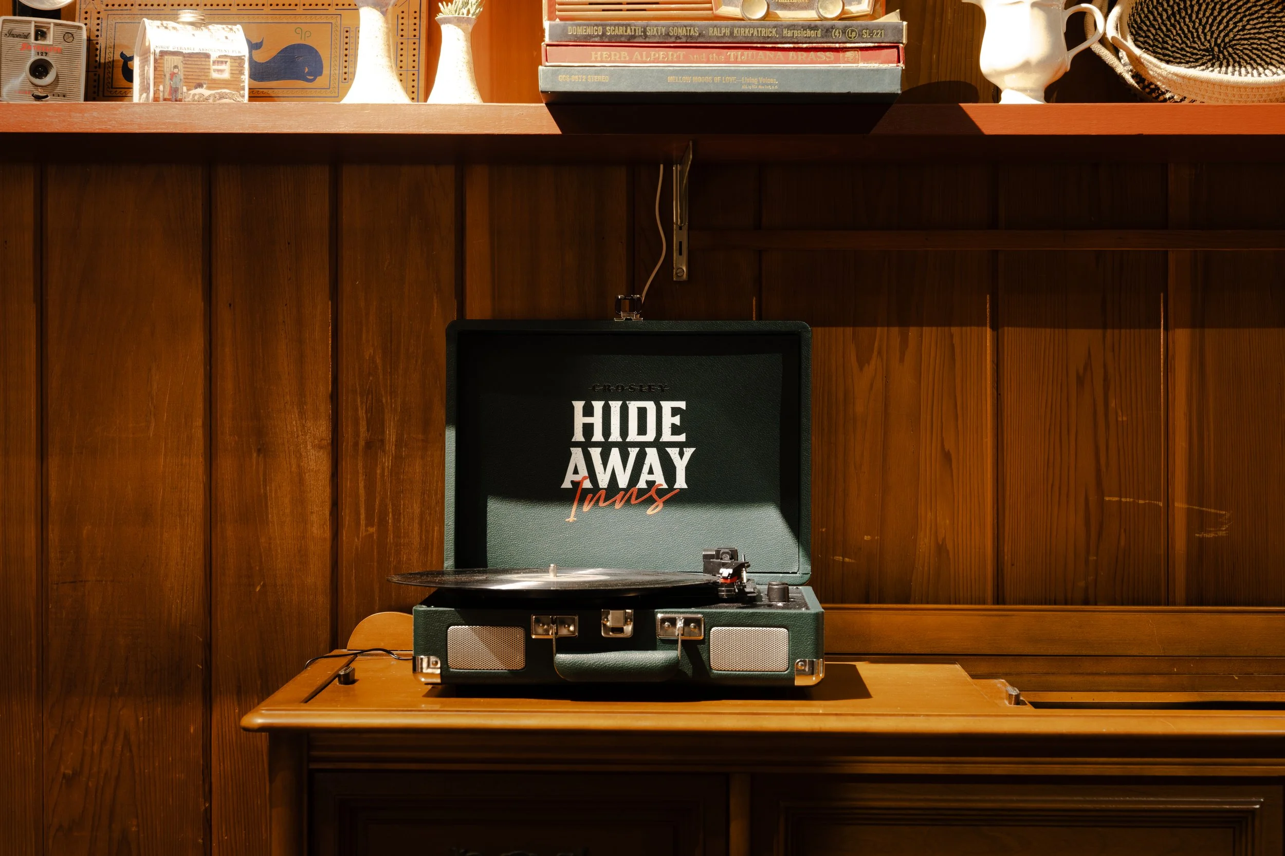 A turntable inside a vintage green case with the words 'HIDE AWAY Inn' on the inside lid. It sits on a wooden surface against a wooden-paneled wall, with shelves above holding books, decorative items, and a camera.