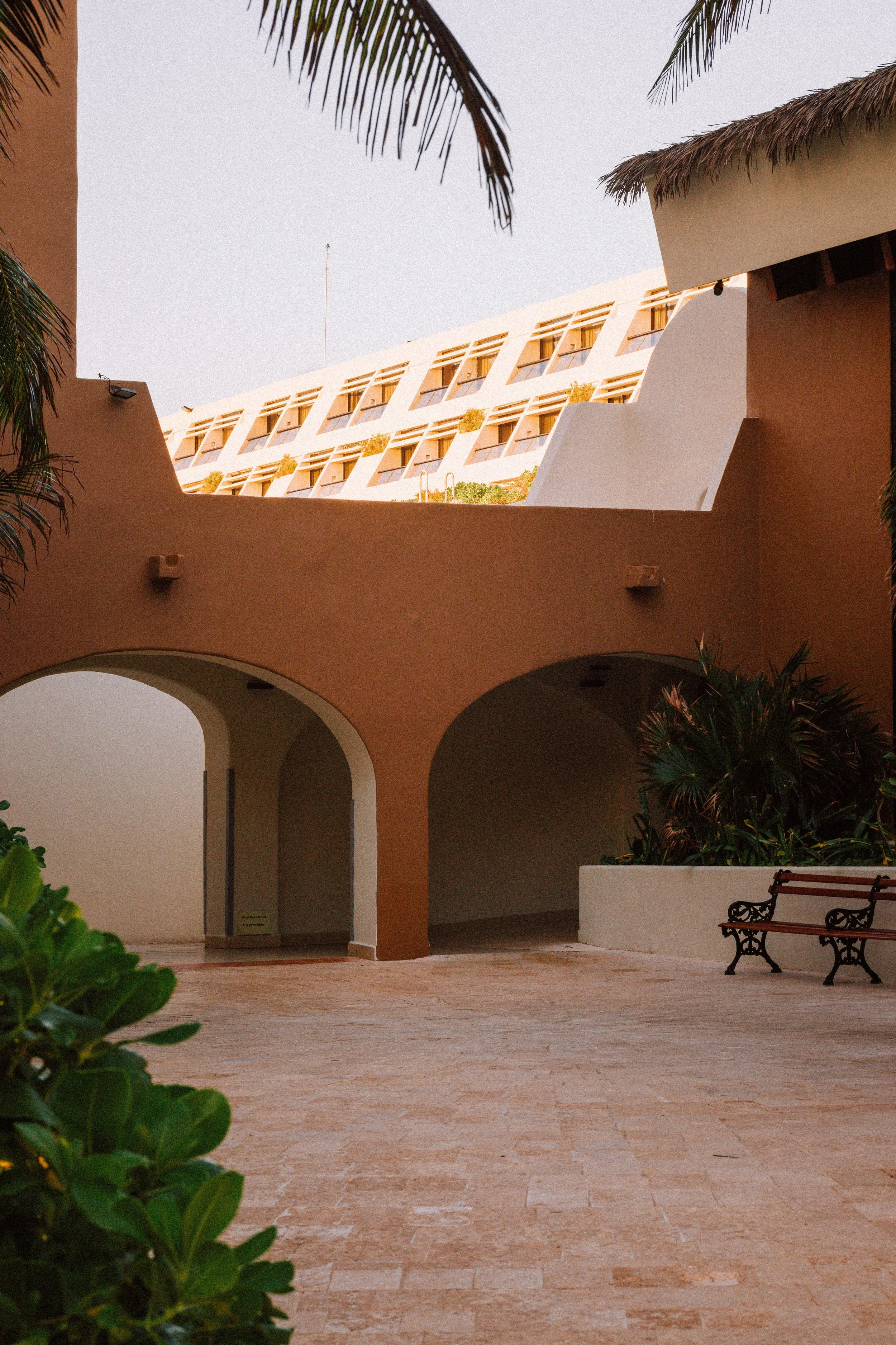 View of a courtyard with arches, a bench, green plants, and a building with balconies viewed through a framed opening.