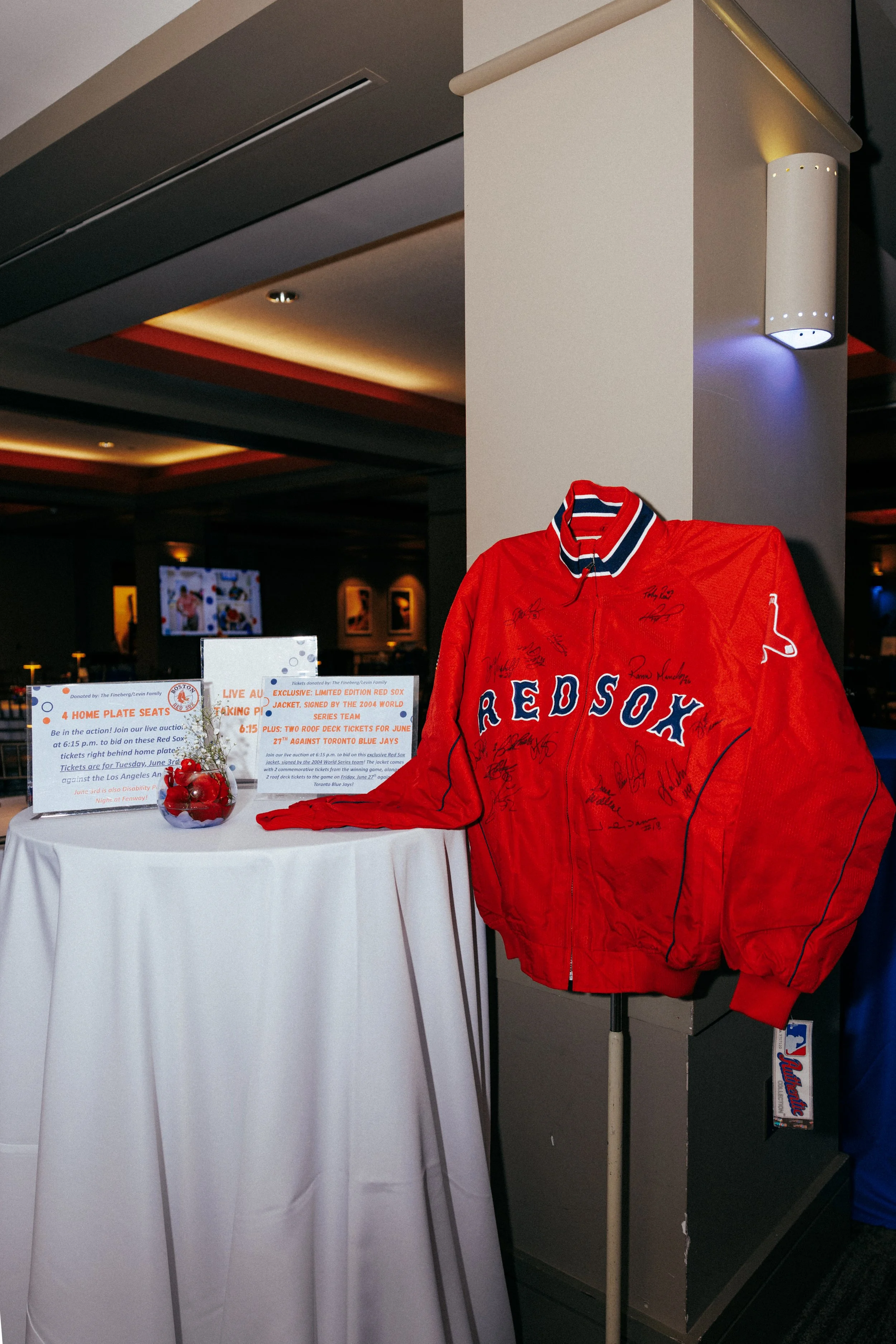 A red Boston Red Sox jacket with several autographs displayed on a mannequin in a dimly lit room, with informational signs and a table nearby.