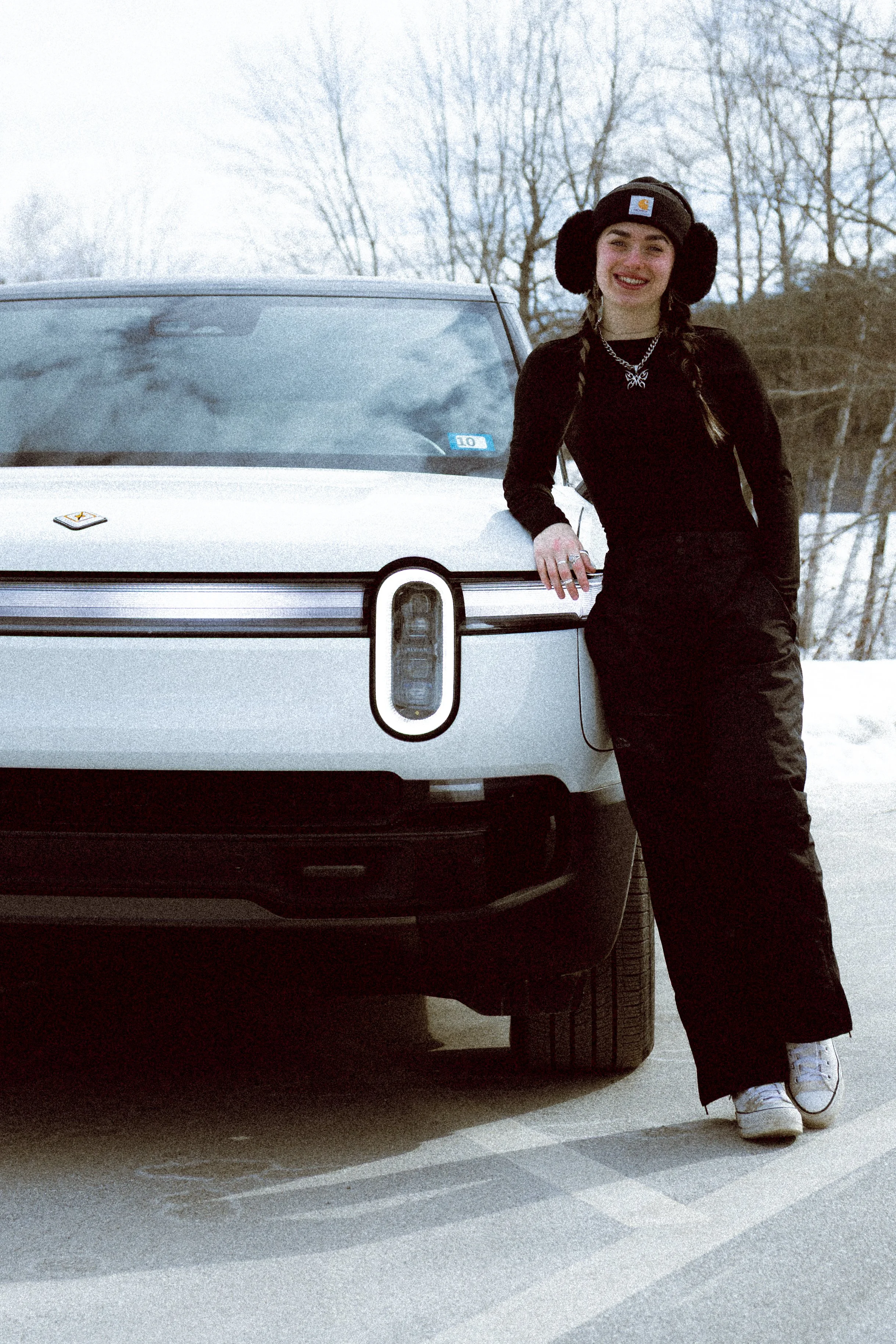 A woman dressed in black winter clothing with earmuffs, standing next to a gray car on a snowy day, smiling at the camera.