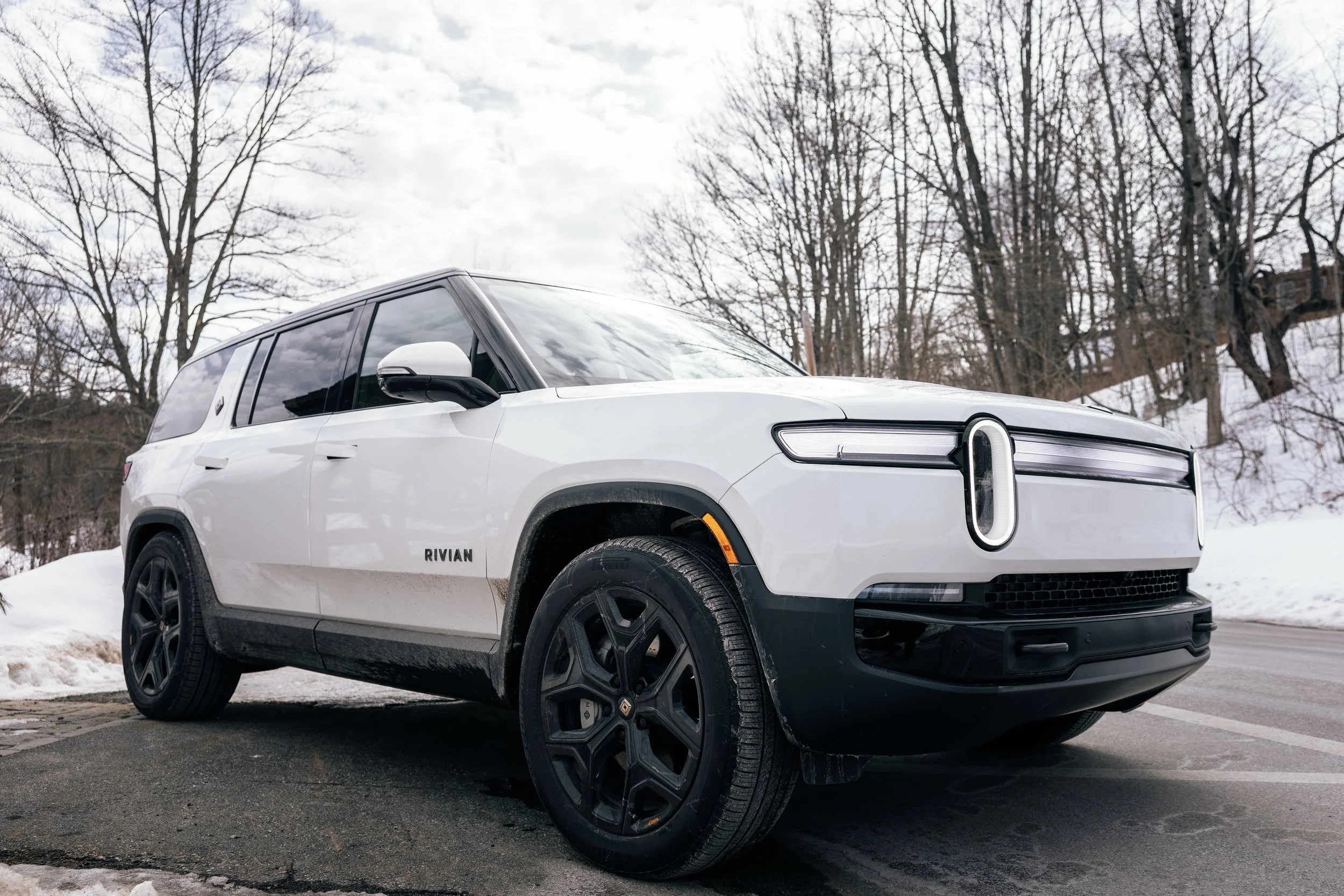White Rivian electric vehicle parked on a winter road with snow and leafless trees in the background