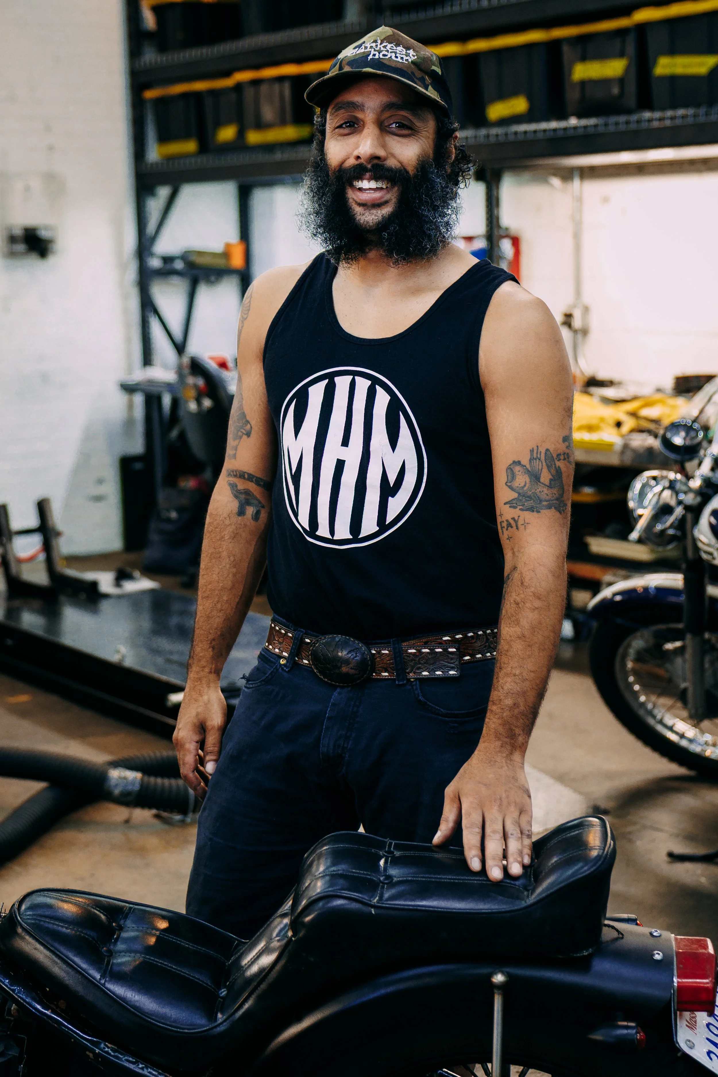 Man with a full beard and tattoos wearing a black tank top and camouflage cap, standing in a garage with motorcycles and tools, resting his hand on a motorcycle seat.