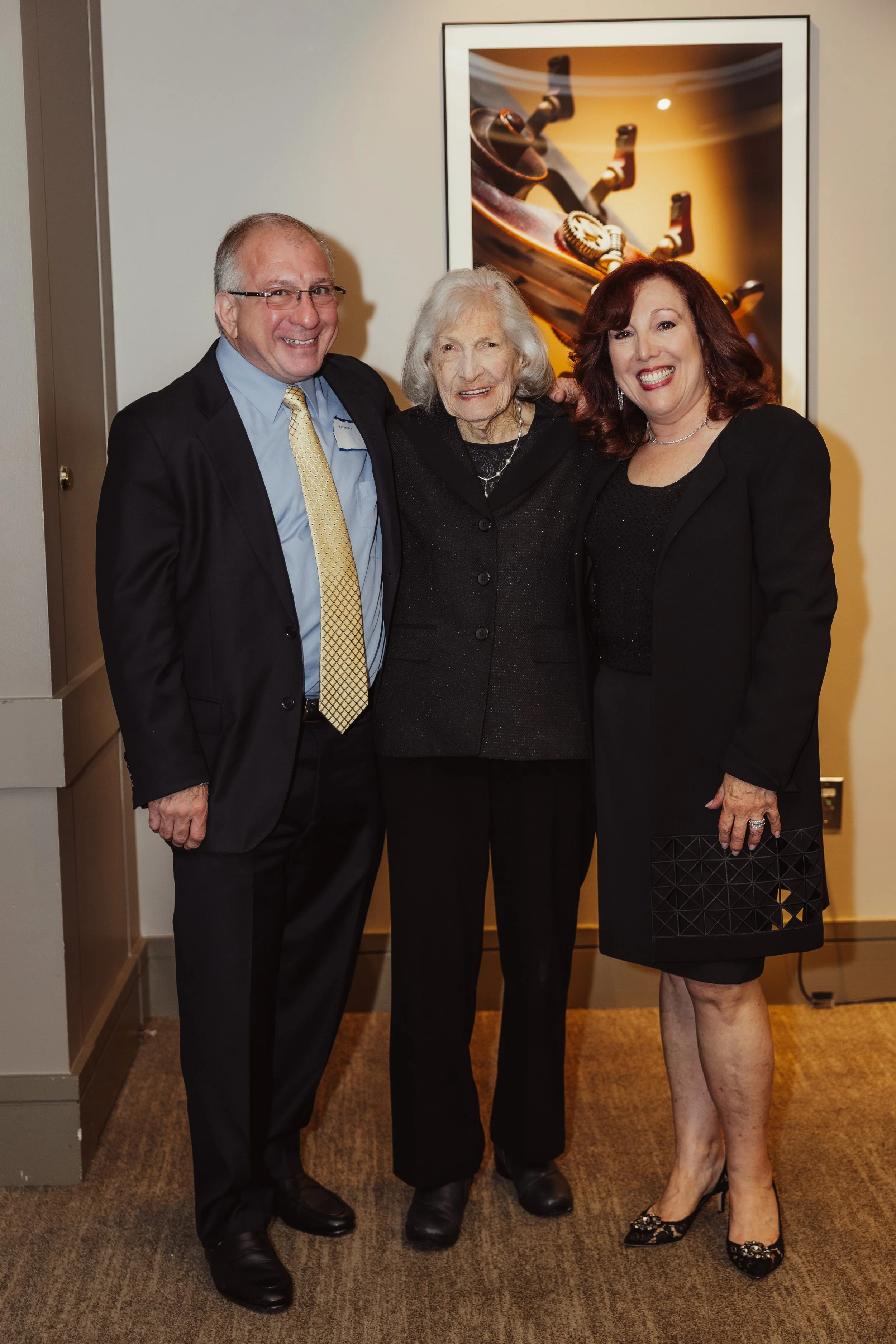 Three people, two women and one man, smiling and standing close together indoors in front of a framed artwork of a tree branch with a lock and chain, dressed in formal attire.