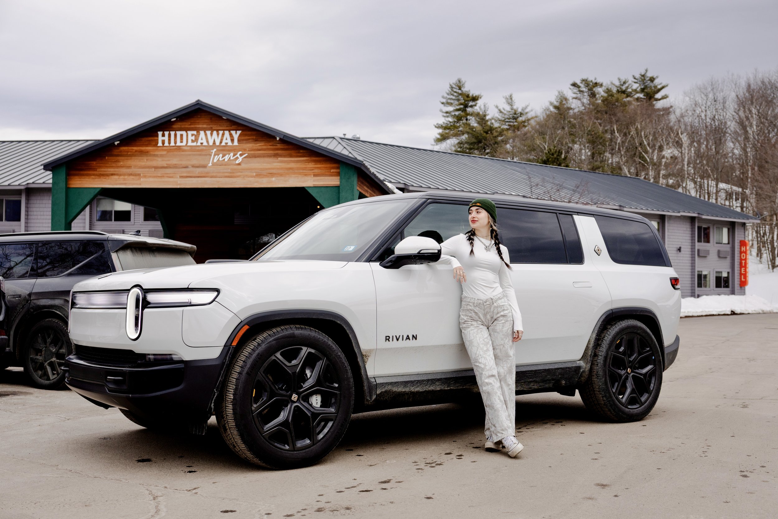 A woman in white and gray standing next to a white Rivian SUV in front of the Hideaway Inn, with a building and snow-covered ground in the background.