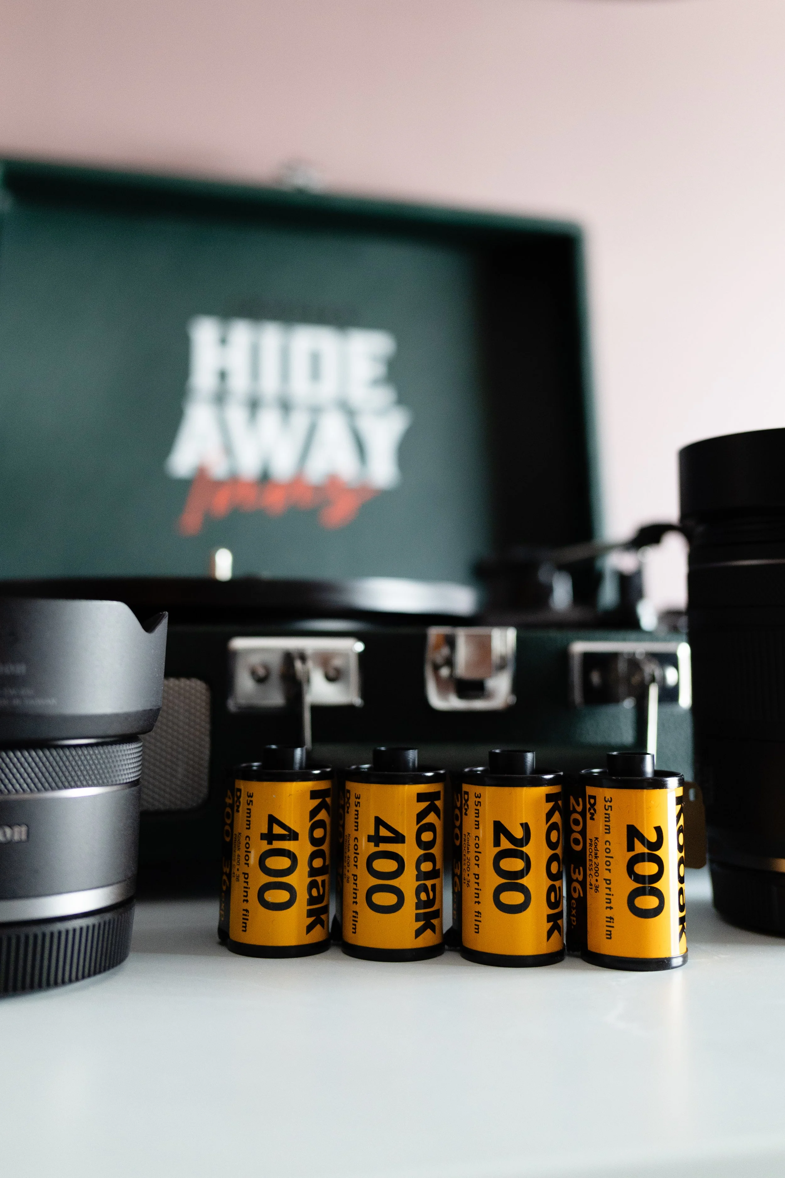 Close-up of four Kodak 200 color film rolls in yellow and black packaging, placed on a white surface with camera lenses nearby. In the background, a record player with a blurred album cover displaying the partially visible text 'HIDE AWAY' and a whit