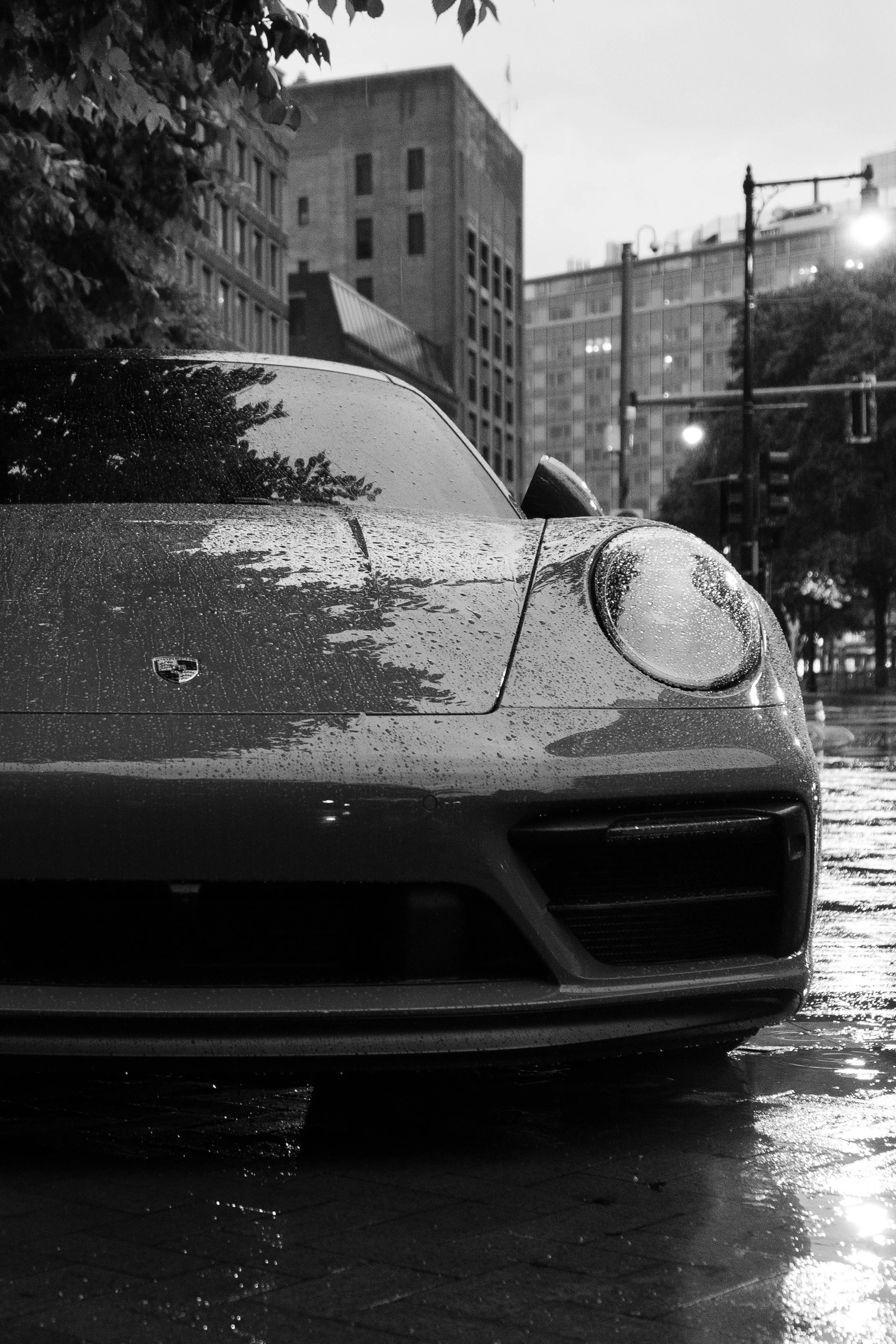 A front view of a Porsche sports car parked on a wet street at night with rain droplets visible on the hood and windshield, in an urban setting with tall buildings and streetlights in the background.